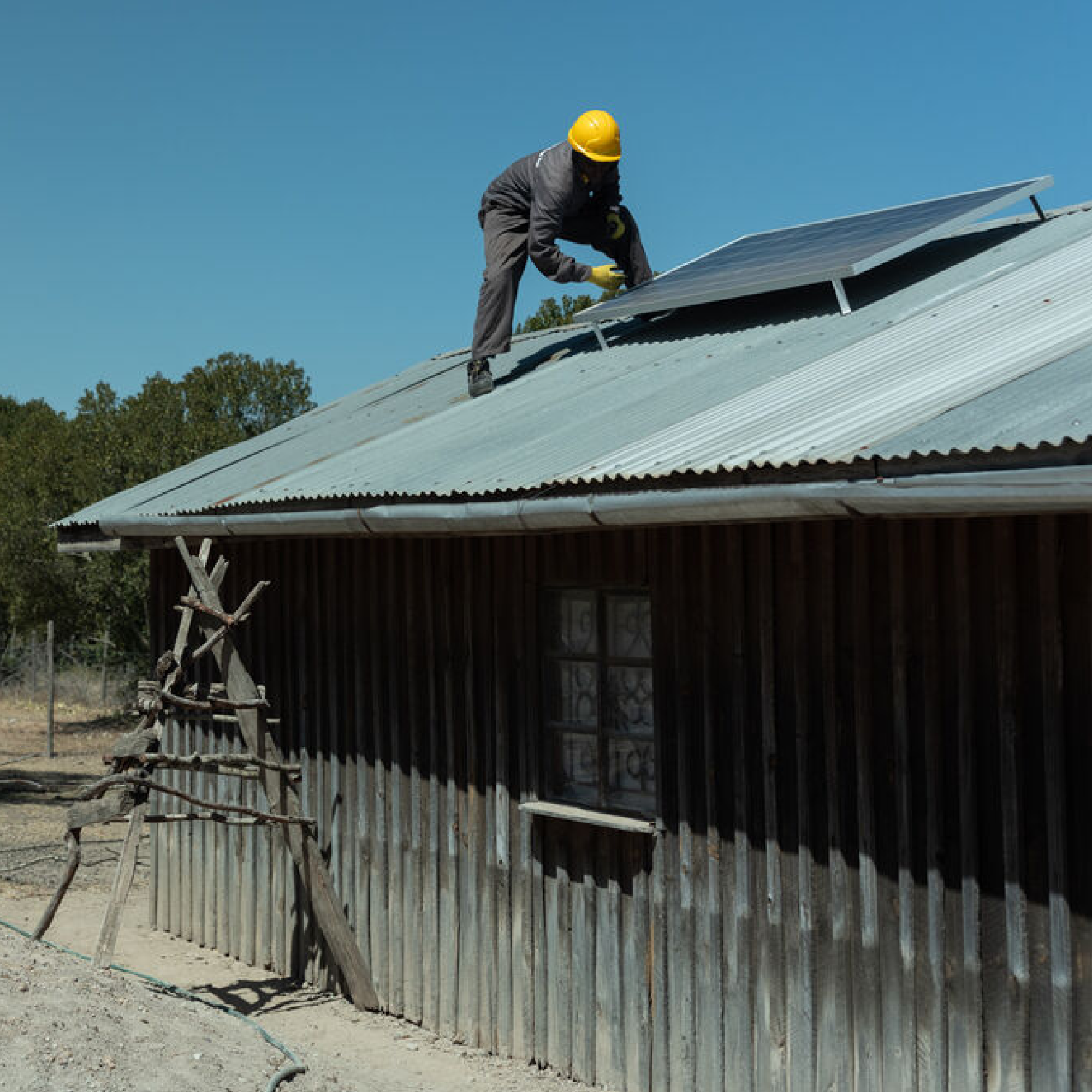 A man wearing a yellow protection hat is on the grey roof of a house, installing a solar panel