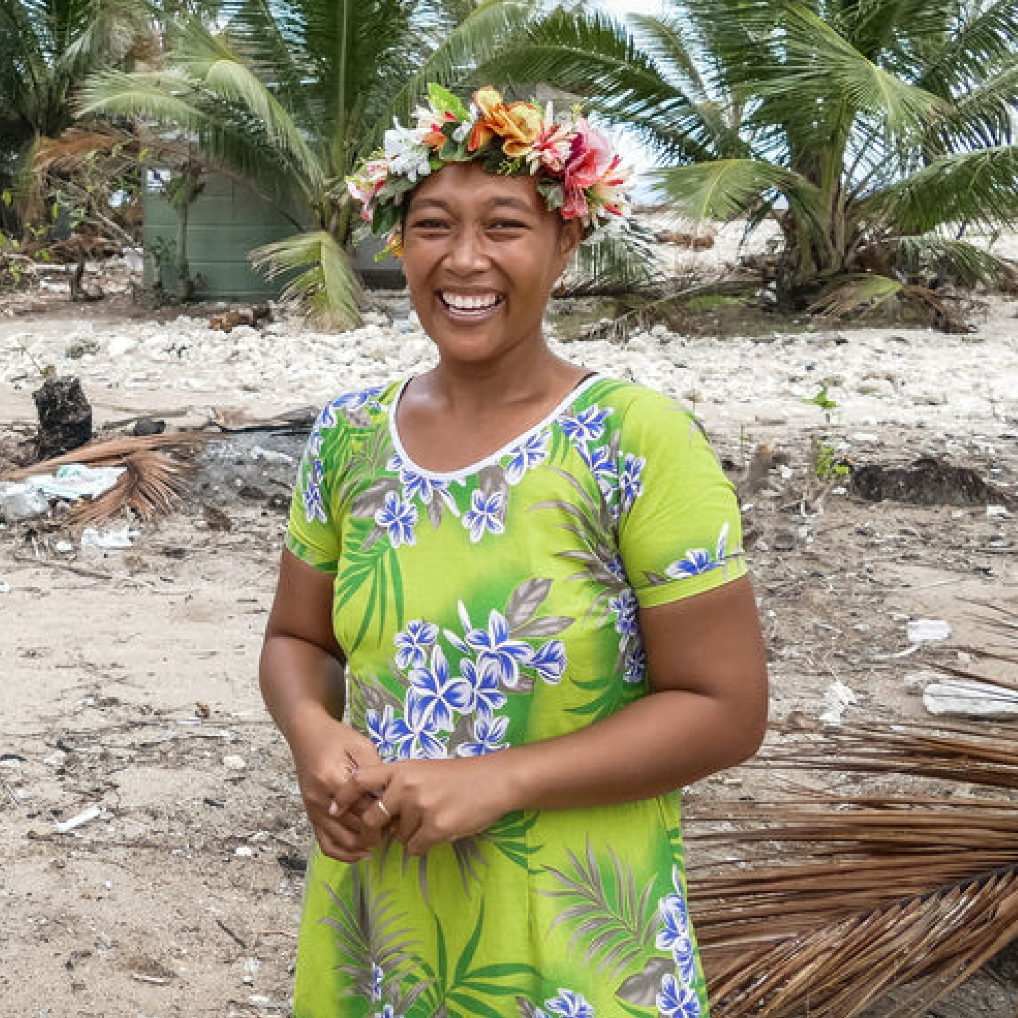 A womand from Tuvalu smiles, wearing flower in her hair, and a light green with purple flowers dress. Behind her, a sand - stony floor and 2 trees