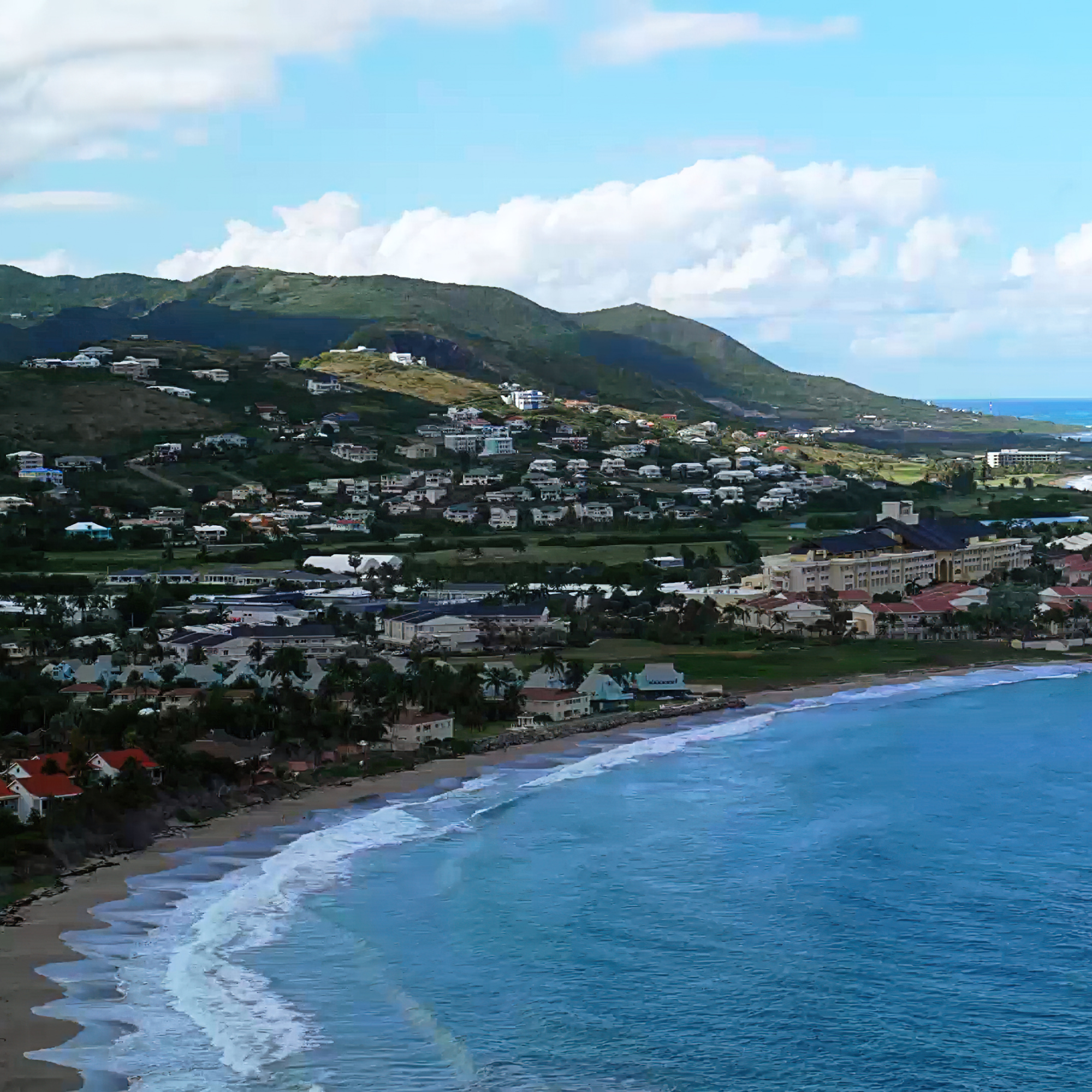View of a mountain with green trees and houses, facing the see. The sky is blue with clouds