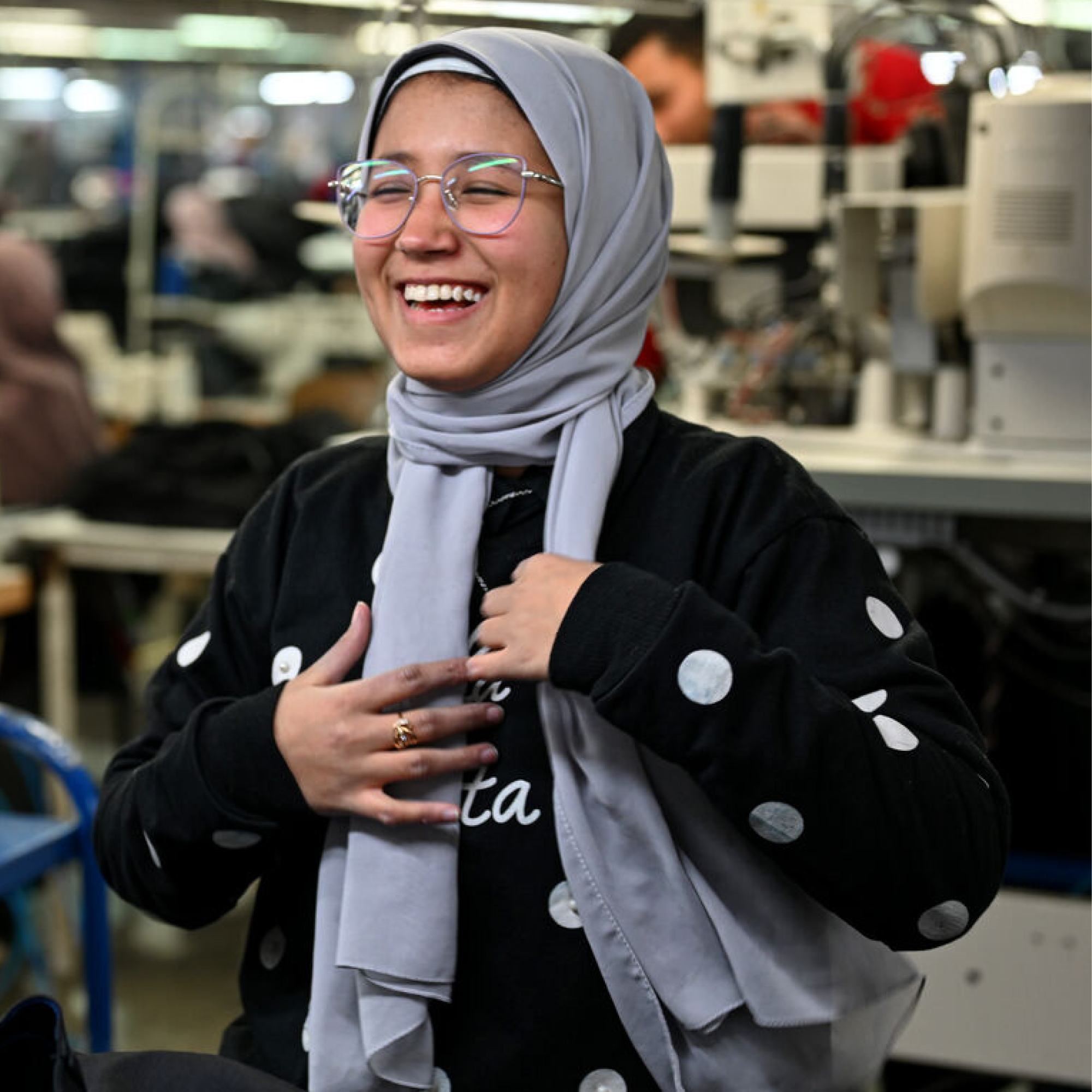 A woman with hair-covered and wearing a black with white dots pull over, and glasses, is smiling. Behind, people and objects