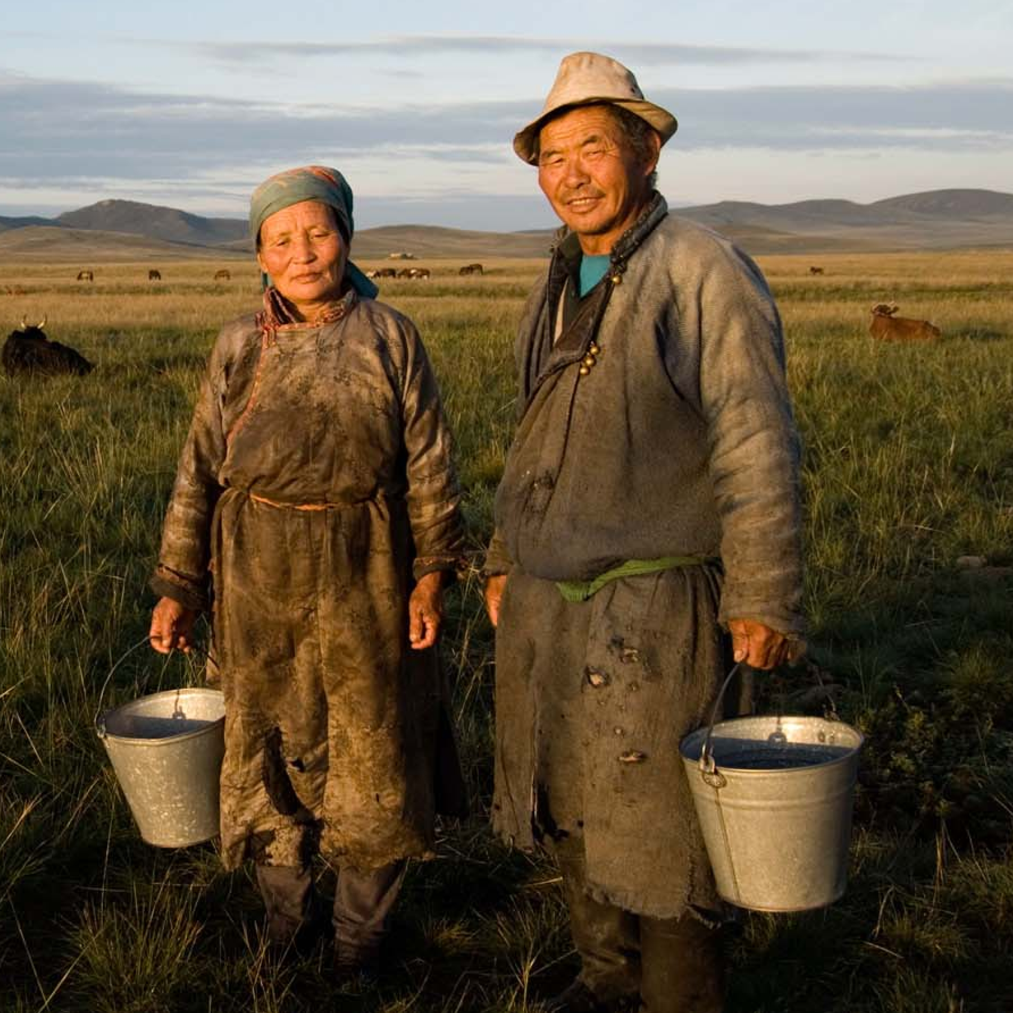 A couple (woman / man) from Mongolia wearing old clothes and boots, holding buckets. He smiles. She looks tired. Backgound of mongolian steppes