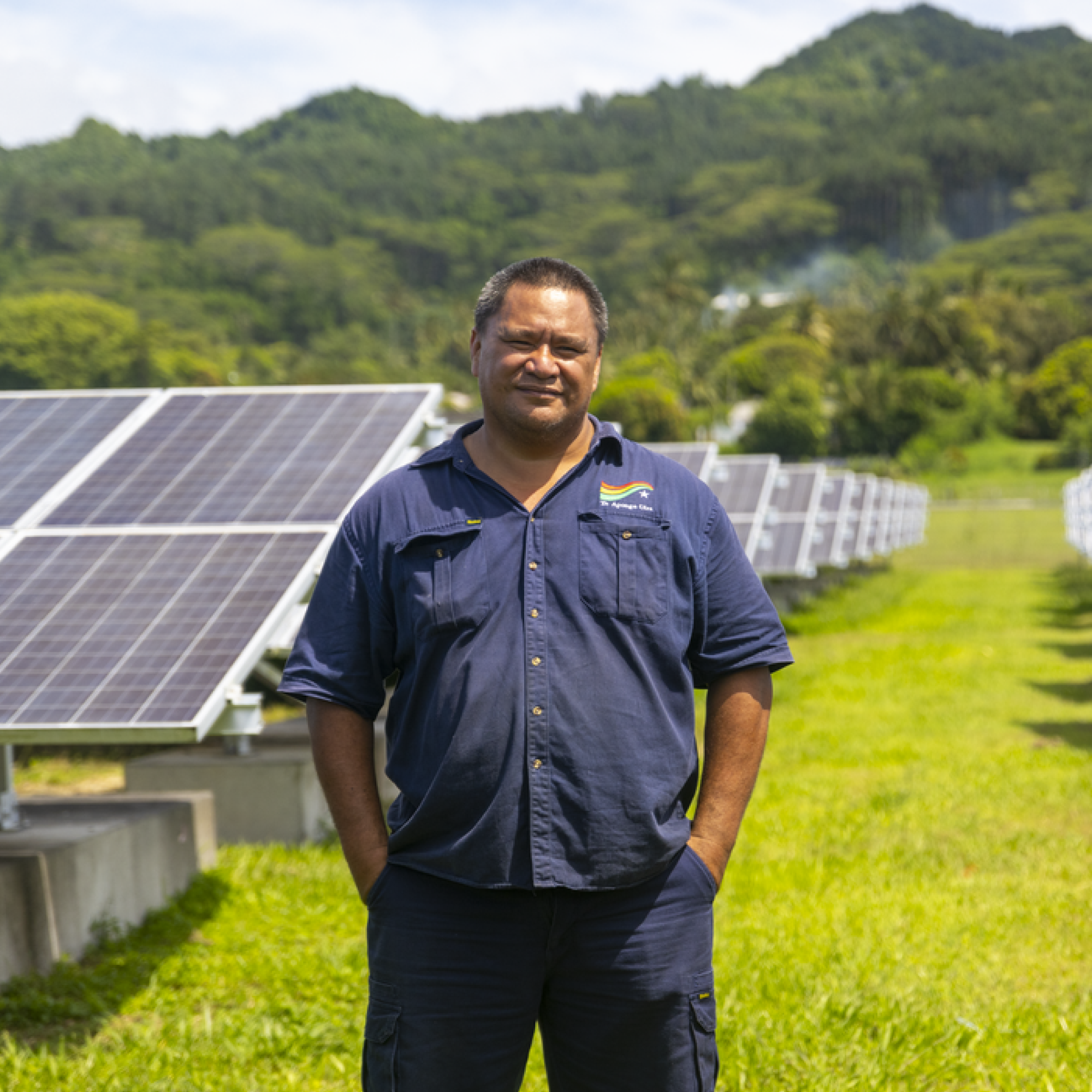 A man wearing dark blue shirt and trousers smiles. Behind solar panels and green grass and mountain.