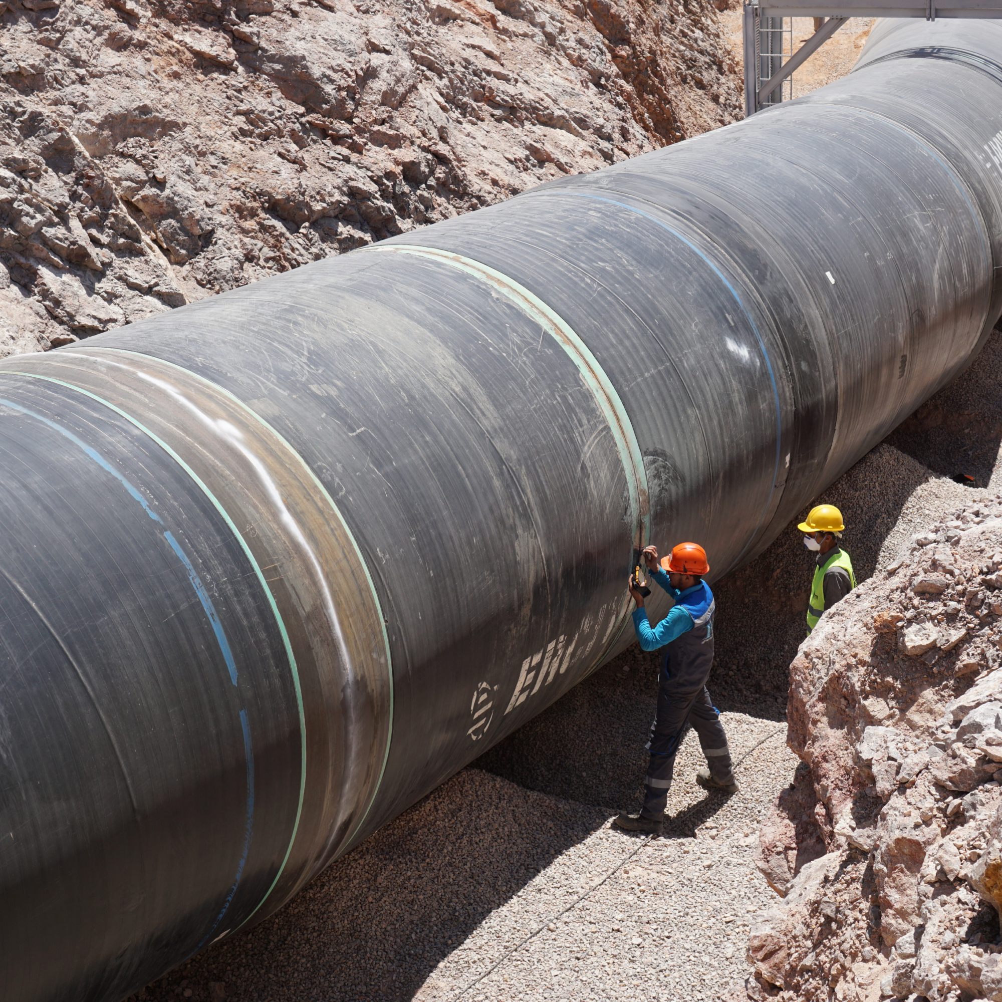 Photograph showing two construction workers inspecting a large black pipeline installed in a rocky trench. Workers wear safety helmets and vests, highlighting scale of pipeline used for transporting fluids or gases in infrastructure projects.