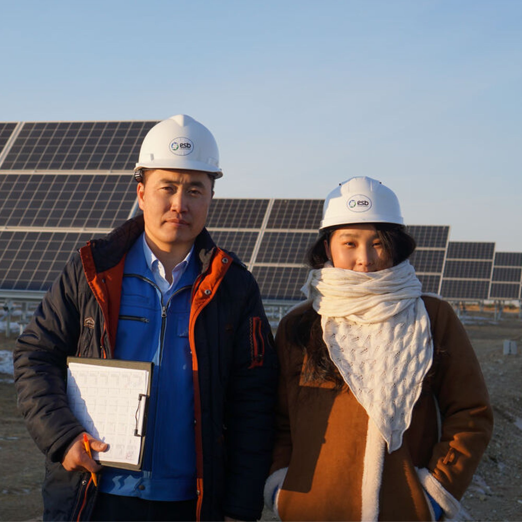 Photograph of two workers from Mongolia - one woman, one man - earing white hard hats and winter clothing standing in front of large solar panel arrays at a solar power facility. One worker holds a clipboard, indicating inspection or monitoring of solar energy equipment during daylight.