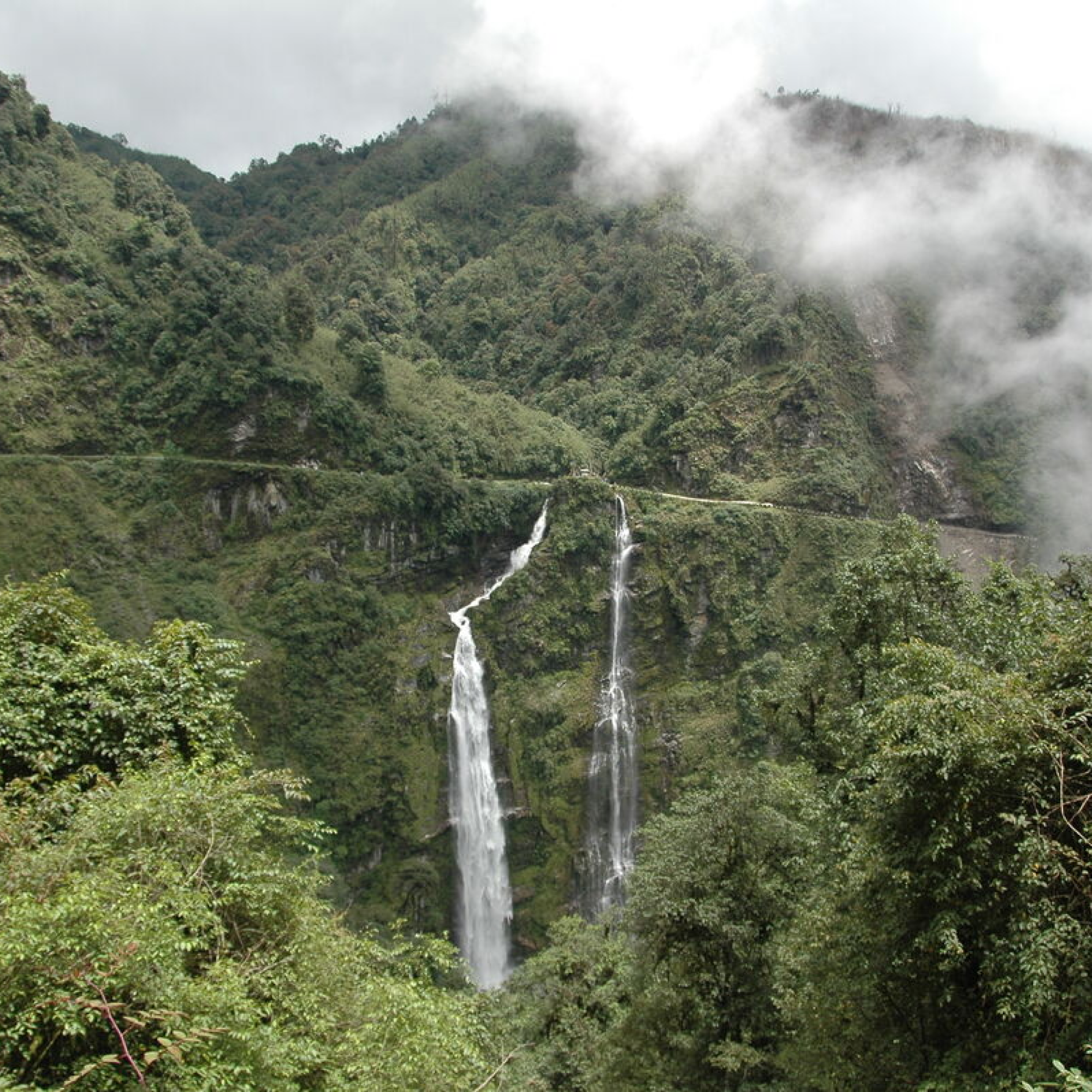 Photograph of a lush green mountainous landscape featuring two prominent waterfalls cascading down steep cliffs. Mist and clouds partially obscure mountain peaks, while dense foliage frames the scene, highlighting natural beauty and serene environment.