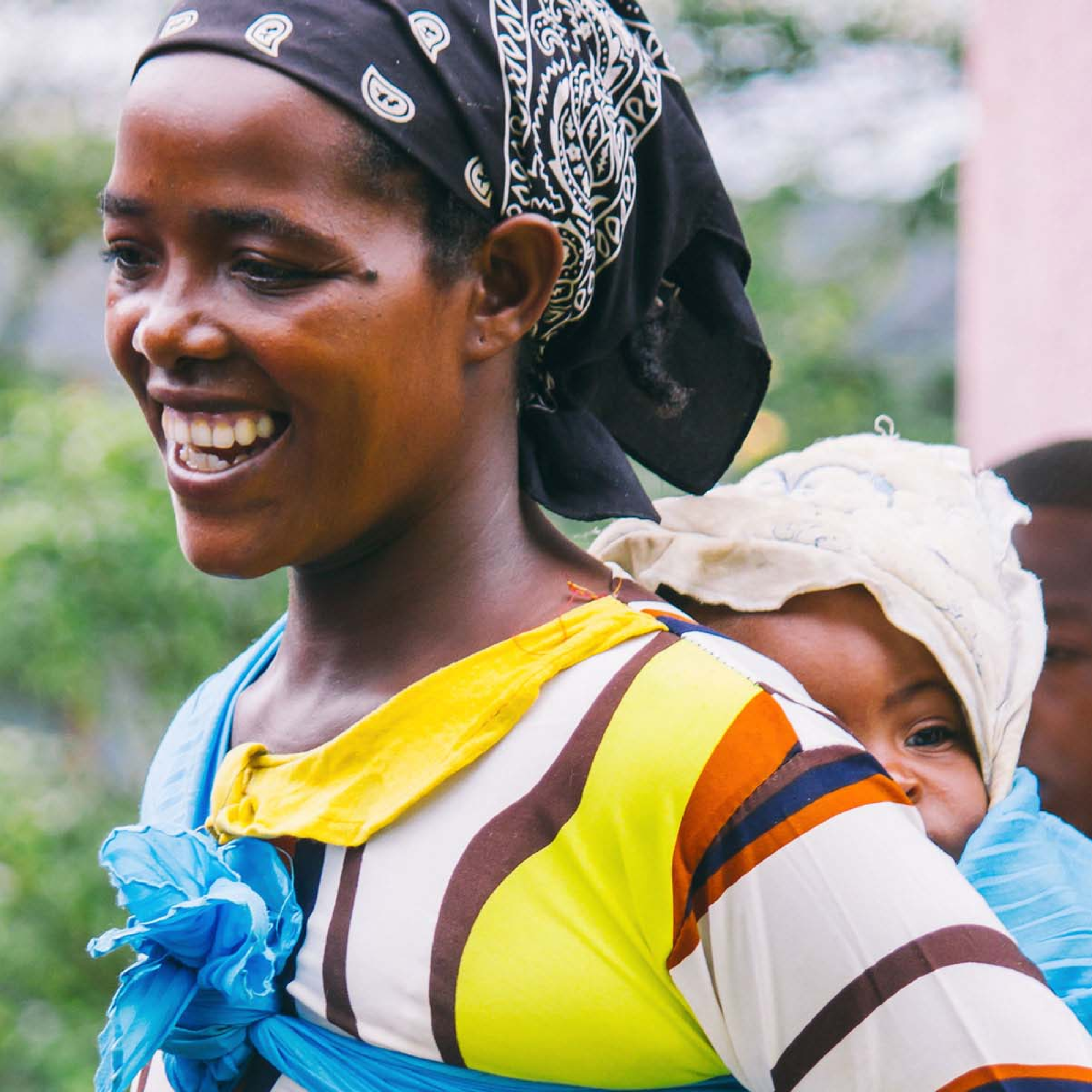Photograph of a smiling black woman wearing a colorful striped shirt with yellow, brown, and white colors, and a black bandana with white patterns on her head. She is carrying a baby wrapped in a light-colored cloth on her back, with a blurred natural background.
