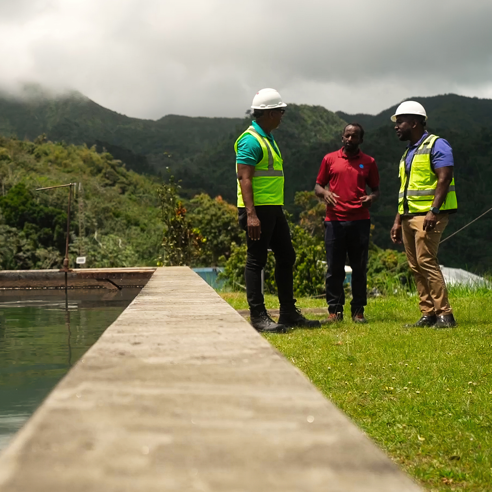 Photograph showing three black men wearing safety helmets and high-visibility vests standing near a concrete water reservoir with green hills and cloudy sky in background. The scene suggests a site inspection or discussion related to water management or environmental work in a mountainous area.