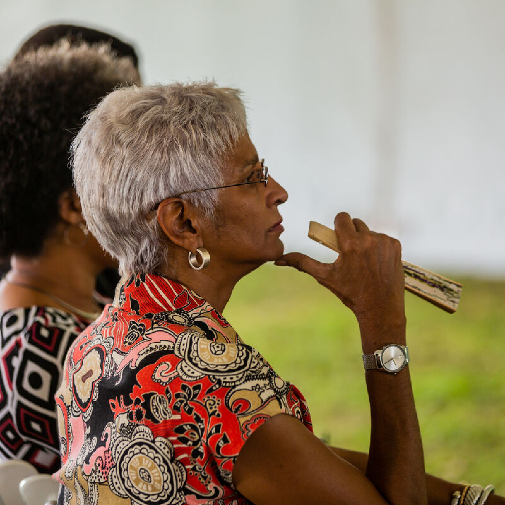 Photograph showing two elderly women seated outdoors, with one woman in the foreground holding a wooden object near her mouth. The scene highlights casual social interaction, with the woman in front wearing a patterned red and black shirt and a wristwatch, set against a blurred green and white background.