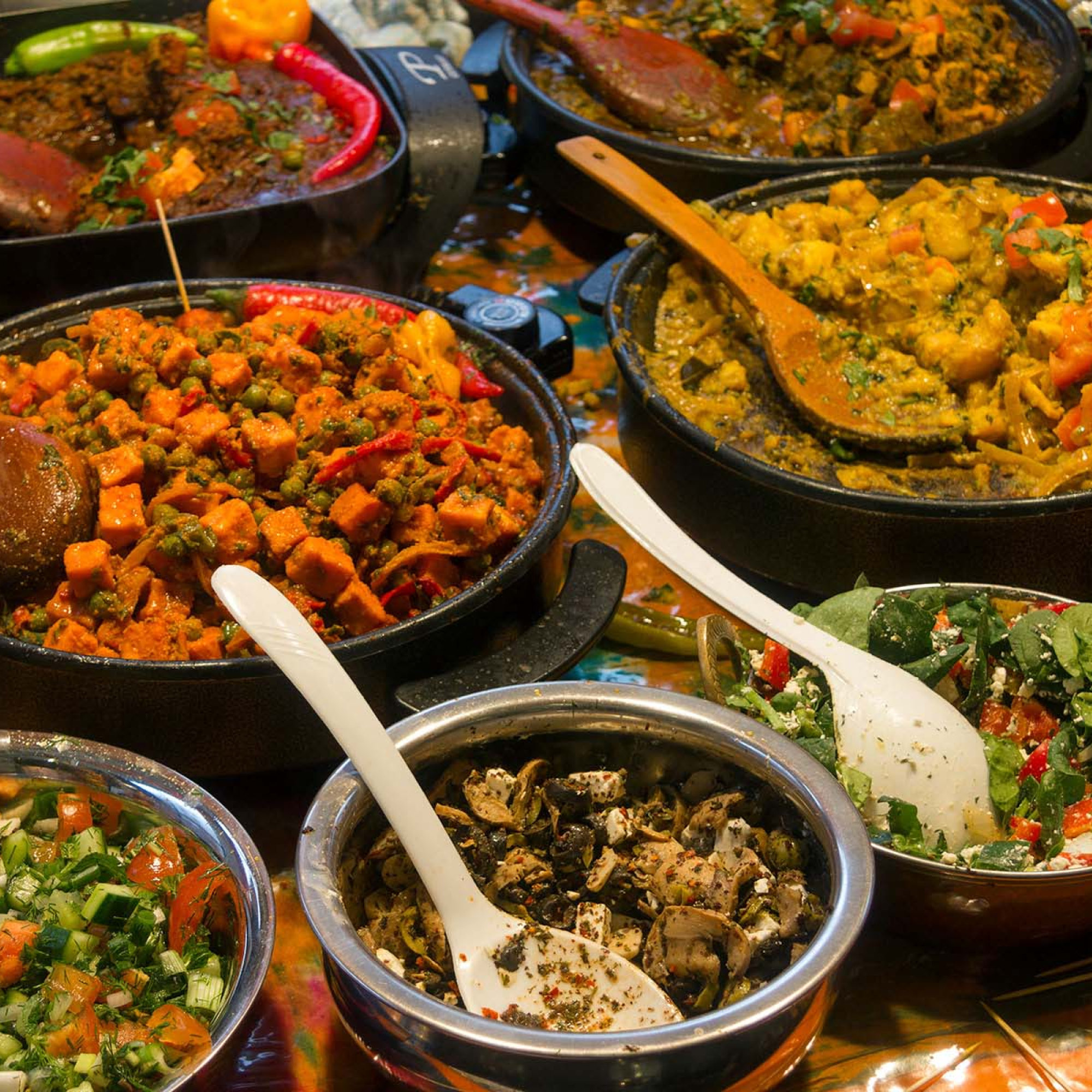 Photograph of a colorful spread of various cooked dishes and salads in bowl and pans, from Bangladesh,showcasing a variety of ingredients like vegetables, herbs, and spices. The setup highlights vibrant colors, textures, and wooden or plastic serving spoons, suggesting a diverse and flavorful meal presentation.