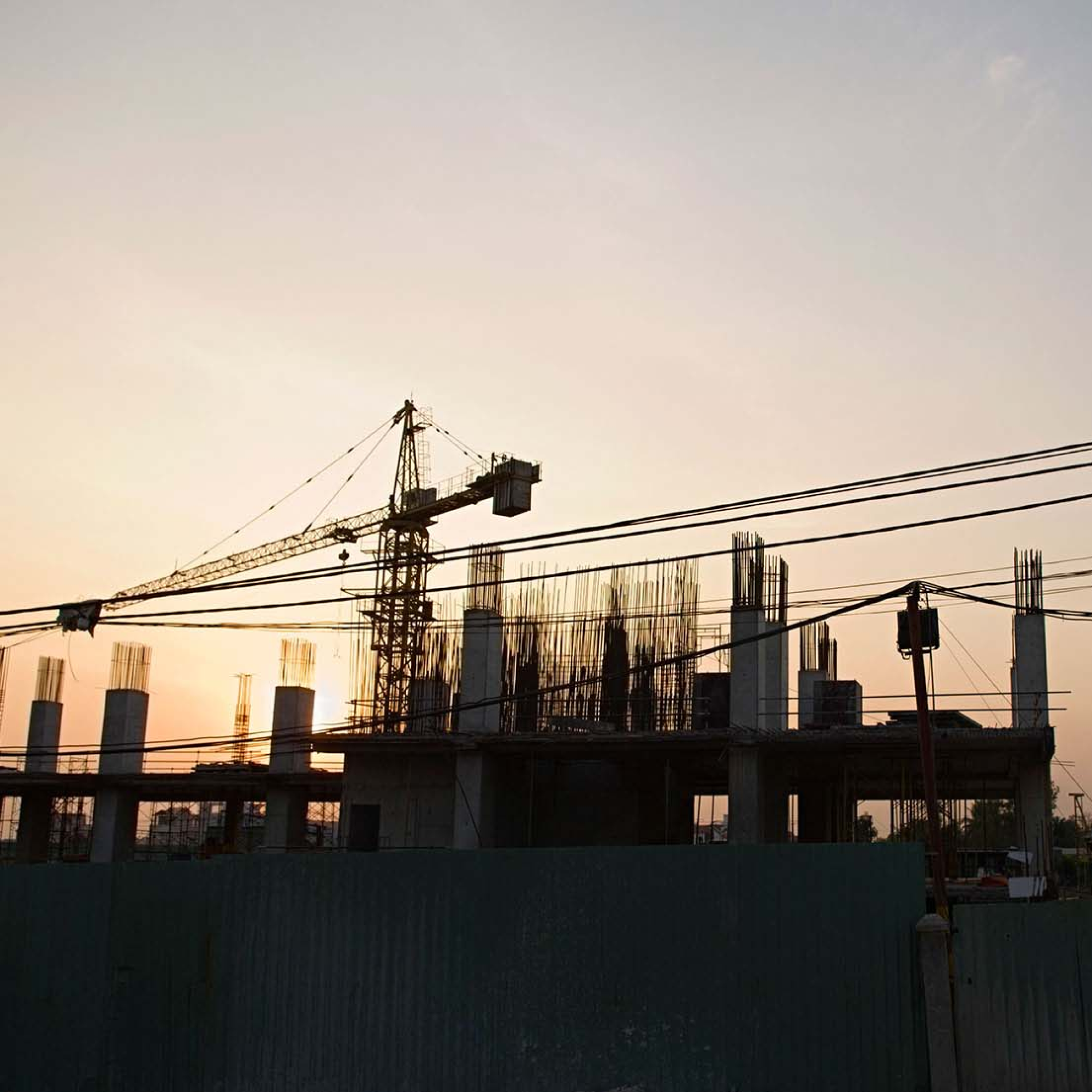 Photograph of a construction site at sunset showing a partially built structure with exposed rebar and concrete columns. A large crane extends over the building, with power lines crossing the foreground against a clear sky.