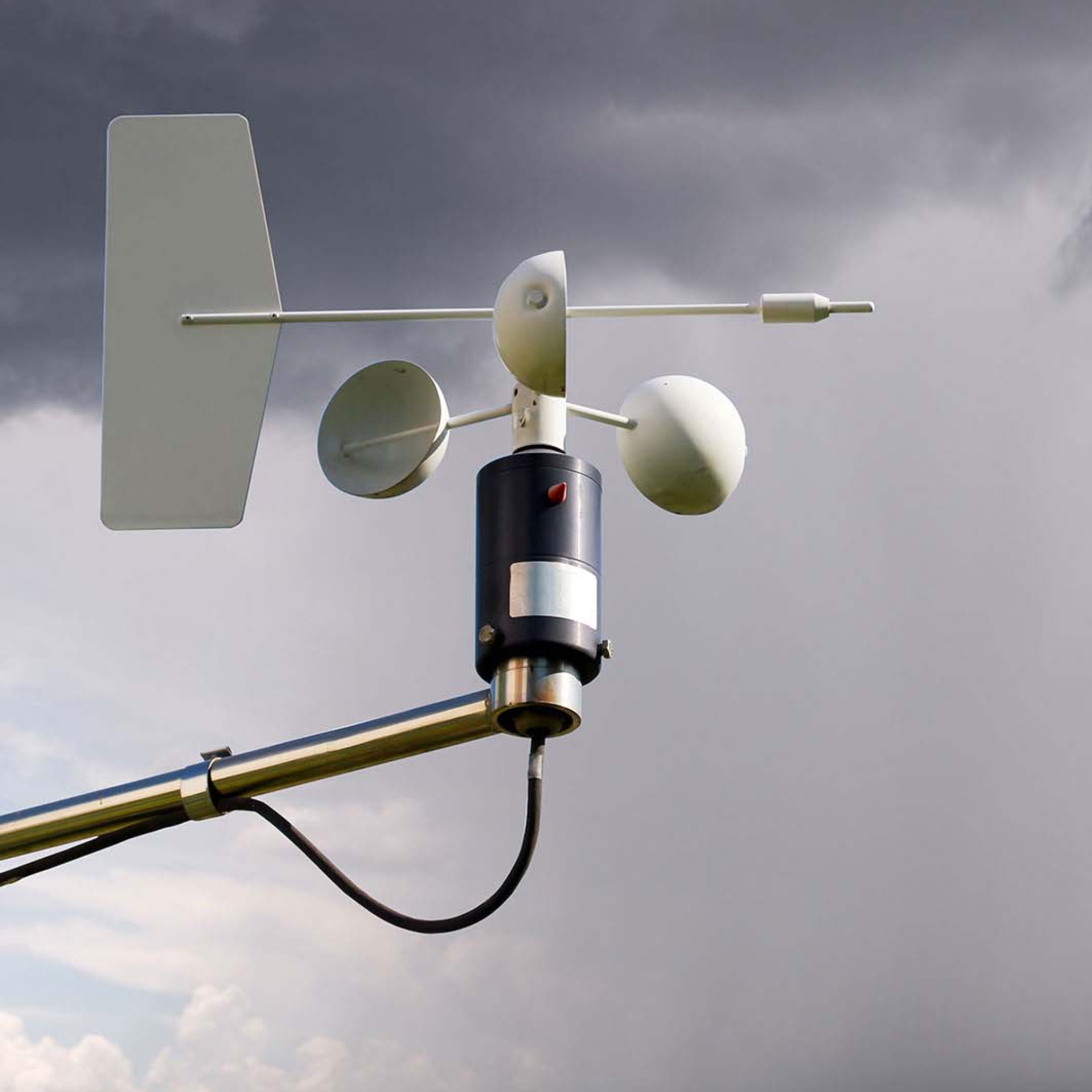 Photograph of an anemometer and wind vane mounted on a metal pole against a cloudy sky, used for measuring wind speed and direction. The device features three white cup-shaped sensors for wind speed and a flat, rectangular tail for wind direction, with a black cylindrical base and attached cable.