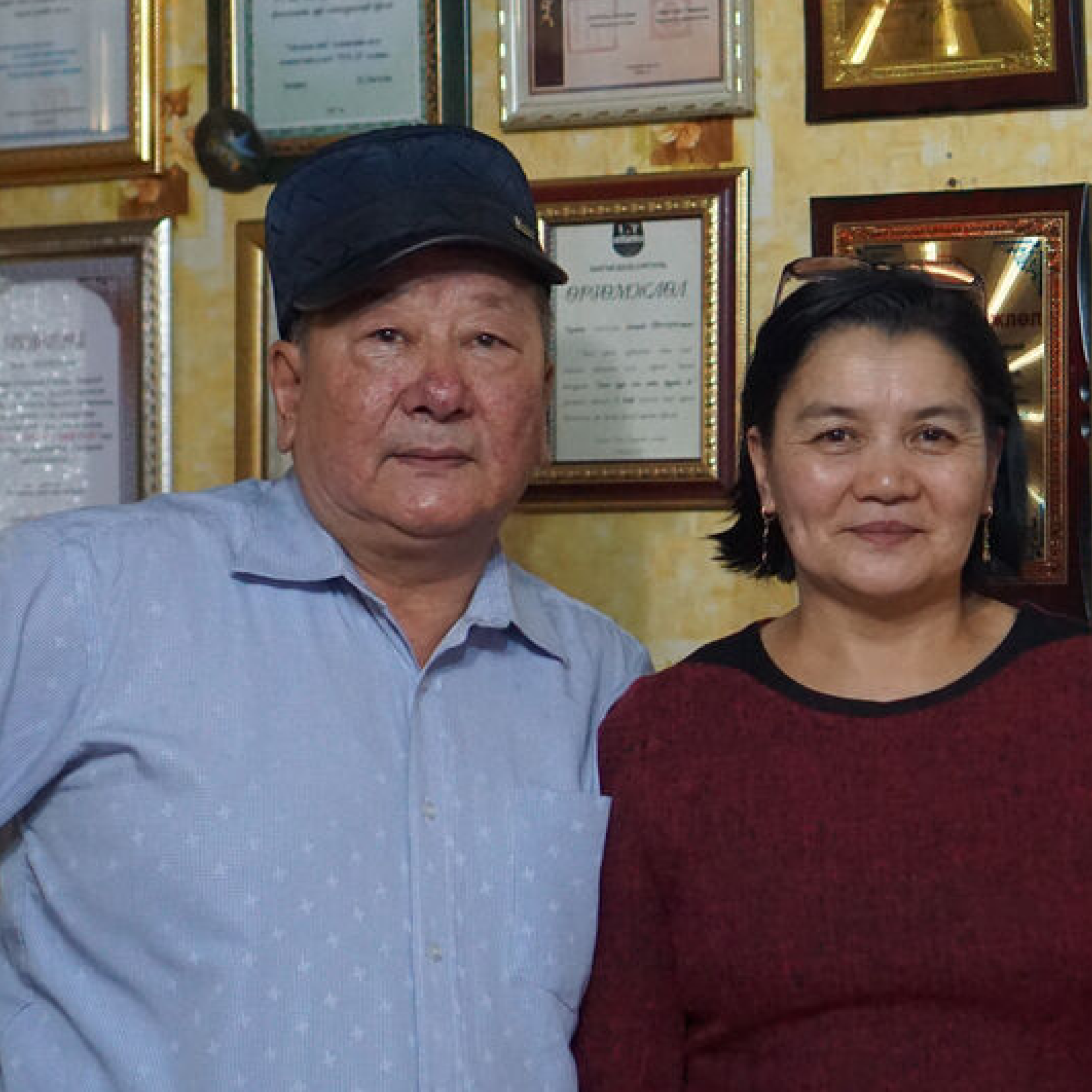 Photograph of two people standing indoors in front of a wall displaying multiple framed certificates and awards. The setting suggests a professional or achievement-oriented environment, with the man wearing a blue shirt and cap and the woman in a maroon top.