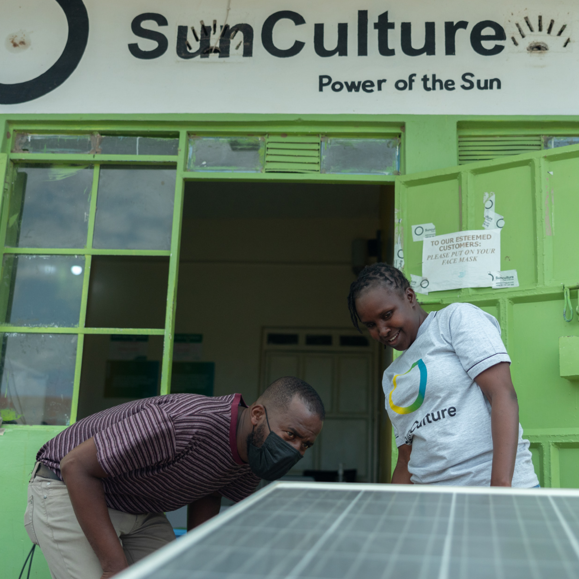 Photograph showing two black people examining a solar panel outside a green building with "SunCulture Power of the Sun" signage above the entrance. One person wears a striped shirt while the other wears a white SunCulture t-shirt, highlighting a focus on solar energy technology and sustainable power solutions.