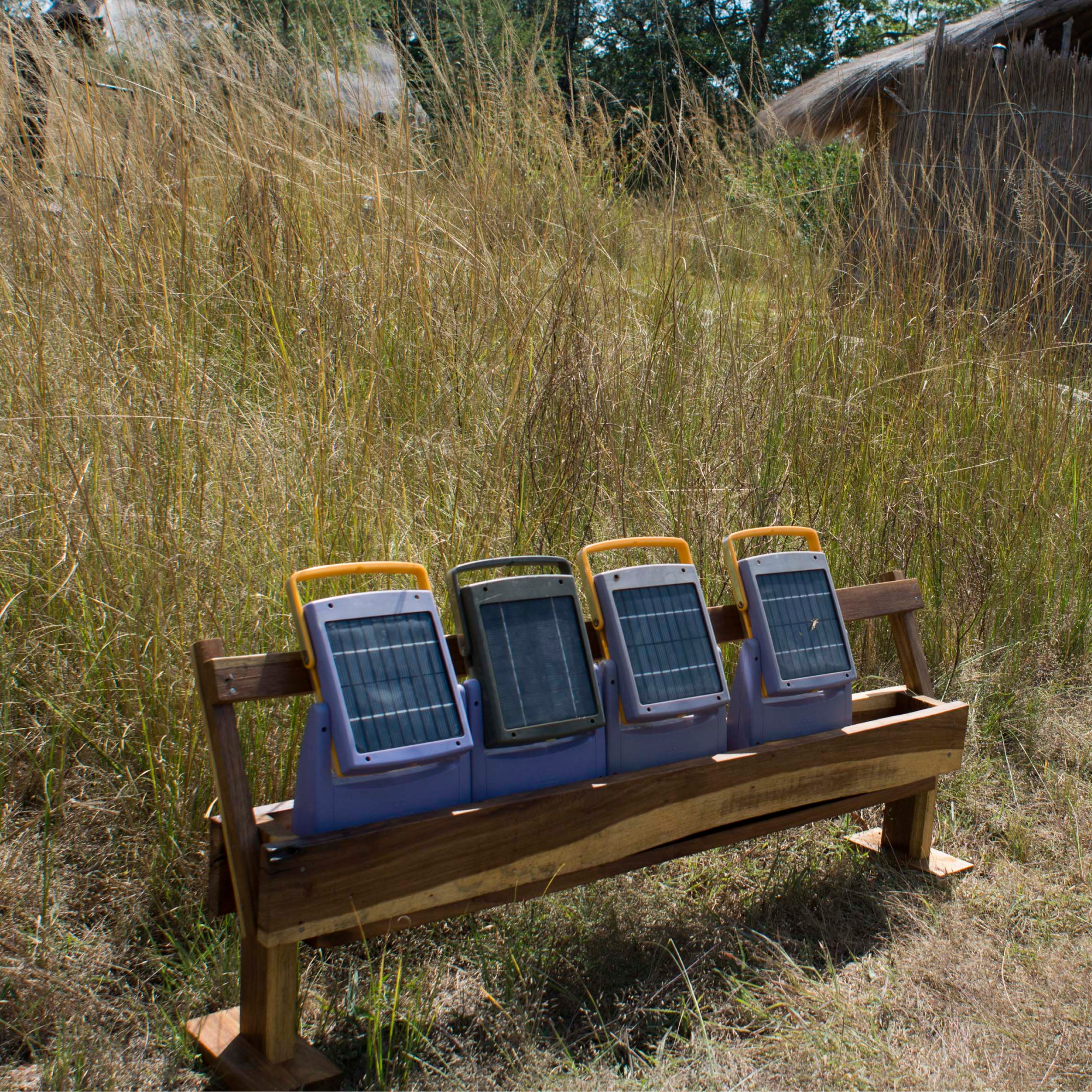 Photograph of four portable solar-powered lanterns with yellow handles placed on a wooden bench in tall dry grass outdoors. Lanterns feature rectangular solar panels on front, suggesting use for off-grid lighting or renewable energy demonstration in rural or natural setting.