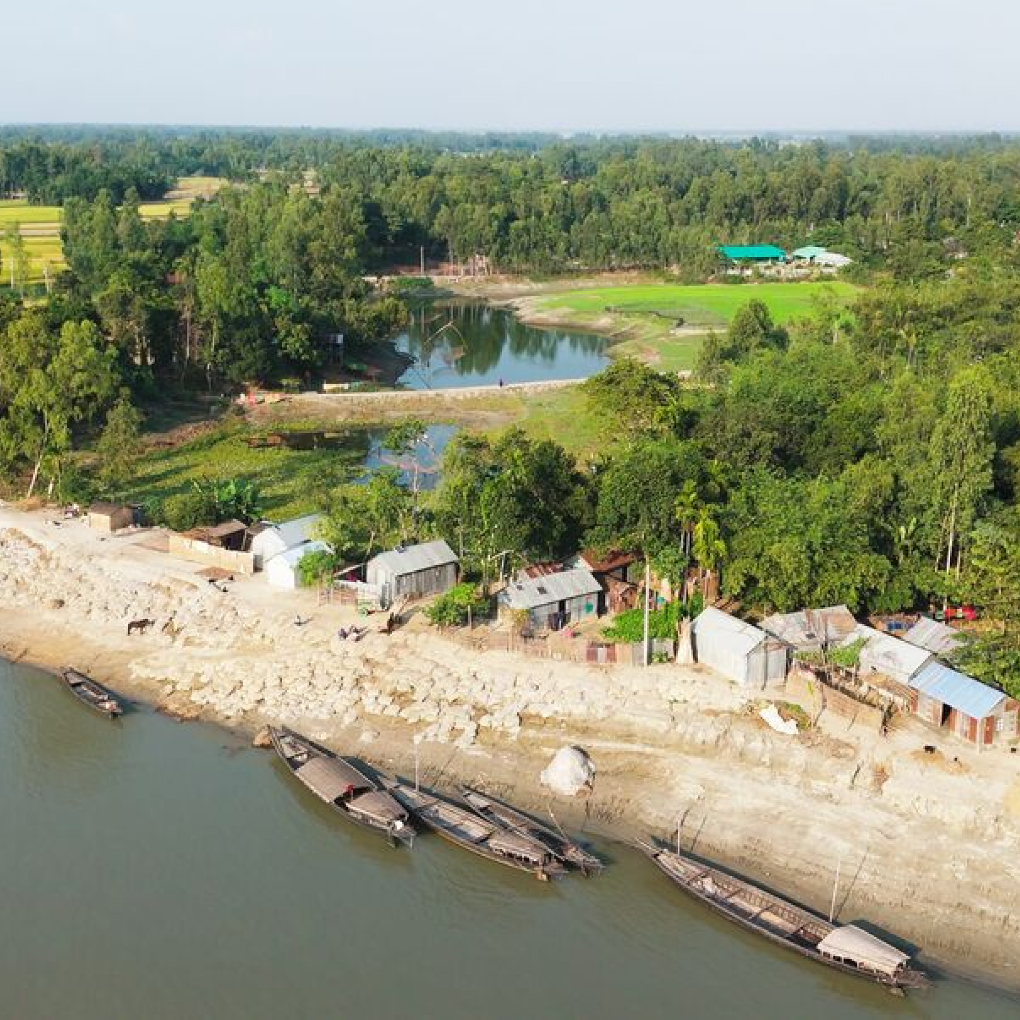 Aerial photograph of a riverside village featuring several wooden boats docked along a sandy shore with small houses and lush green trees in the background. The scene highlights rural life with a calm river, dense forest, and agricultural fields extending into the distance.