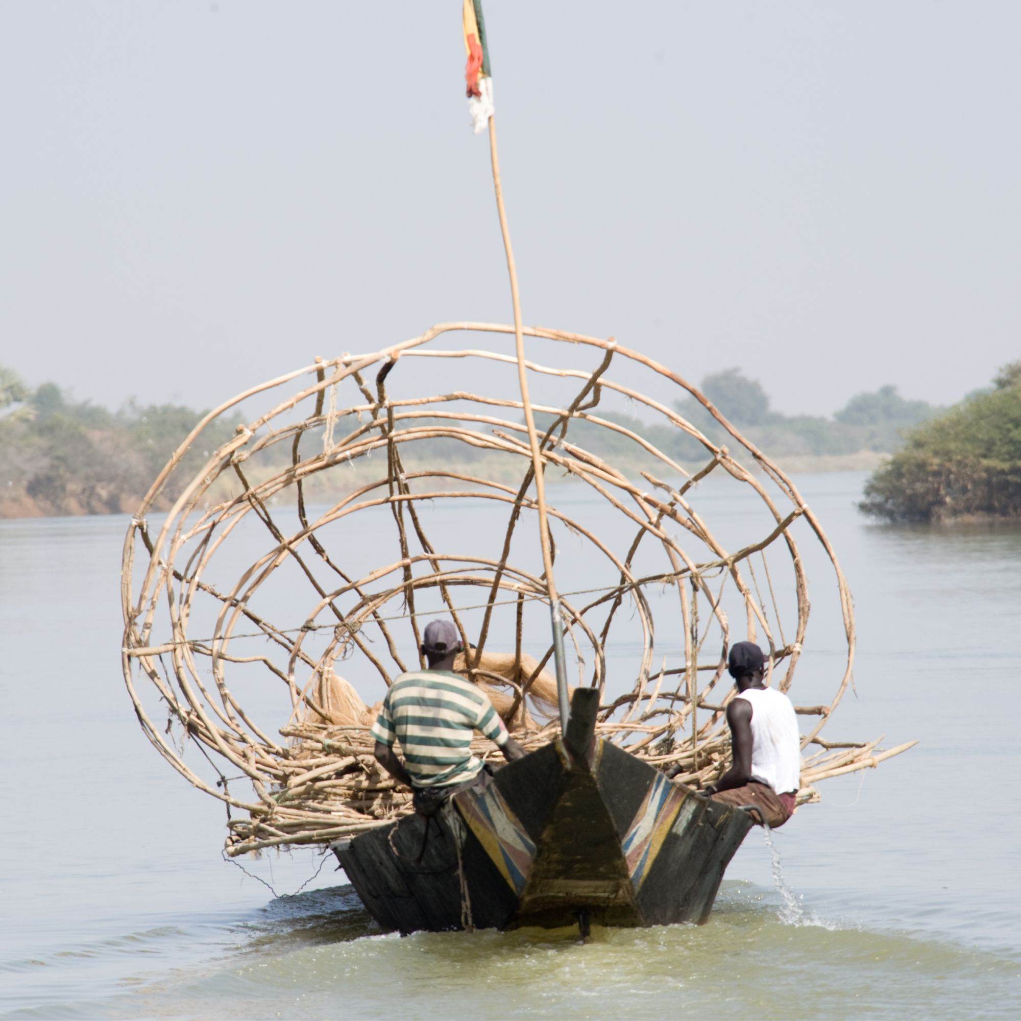 Photograph of two men on a wooden boat carrying a large circular fishing trap made of woven sticks, floating on a calm river. Background includes distant trees and hazy sky, highlighting traditional fishing methods in a rural waterway setting.