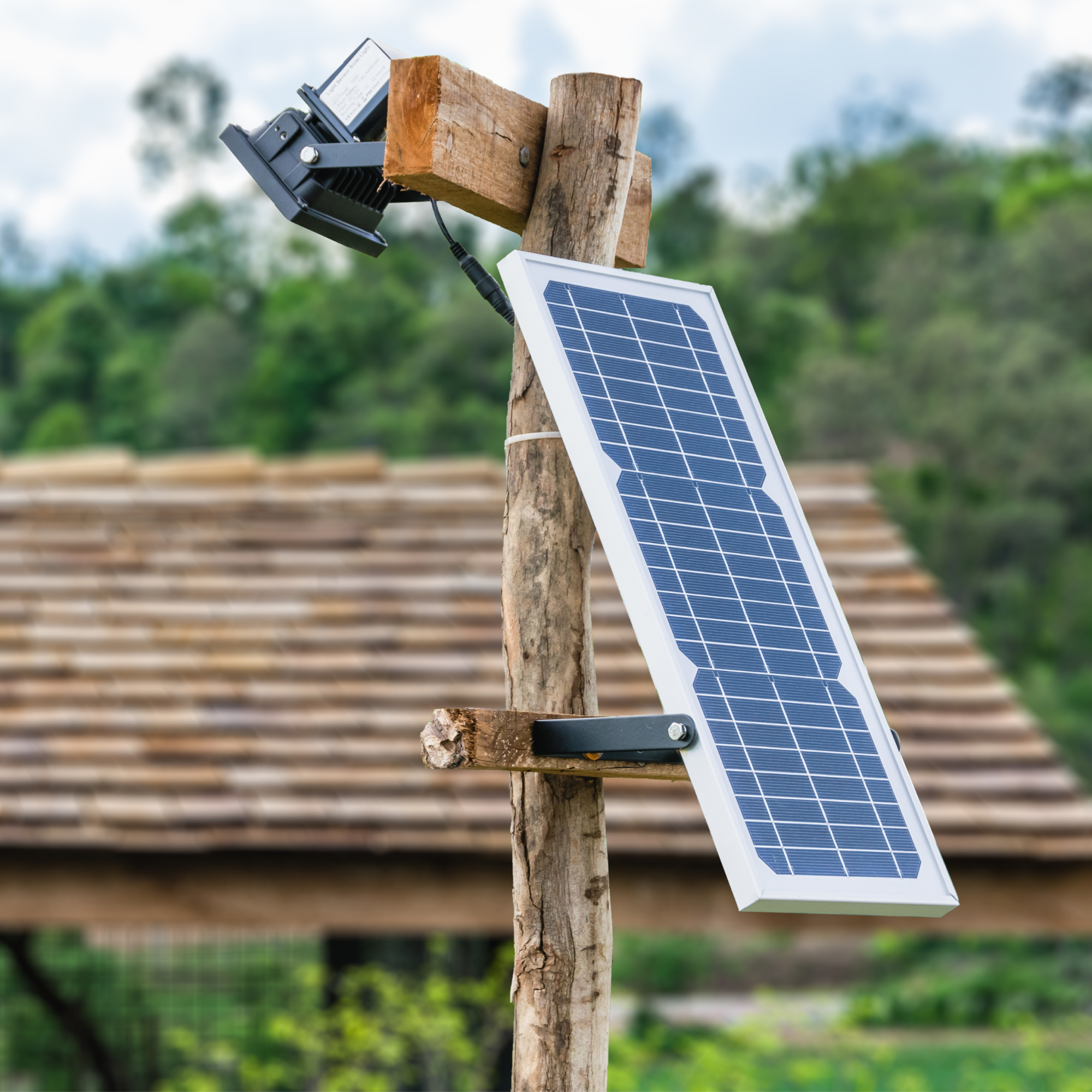 Photograph of a small solar panel mounted on a wooden post with a light fixture attached above it, set against a blurred background of greenery and a rustic roof. The setup highlights a simple off-grid solar power solution likely used for outdoor lighting or small-scale energy needs.