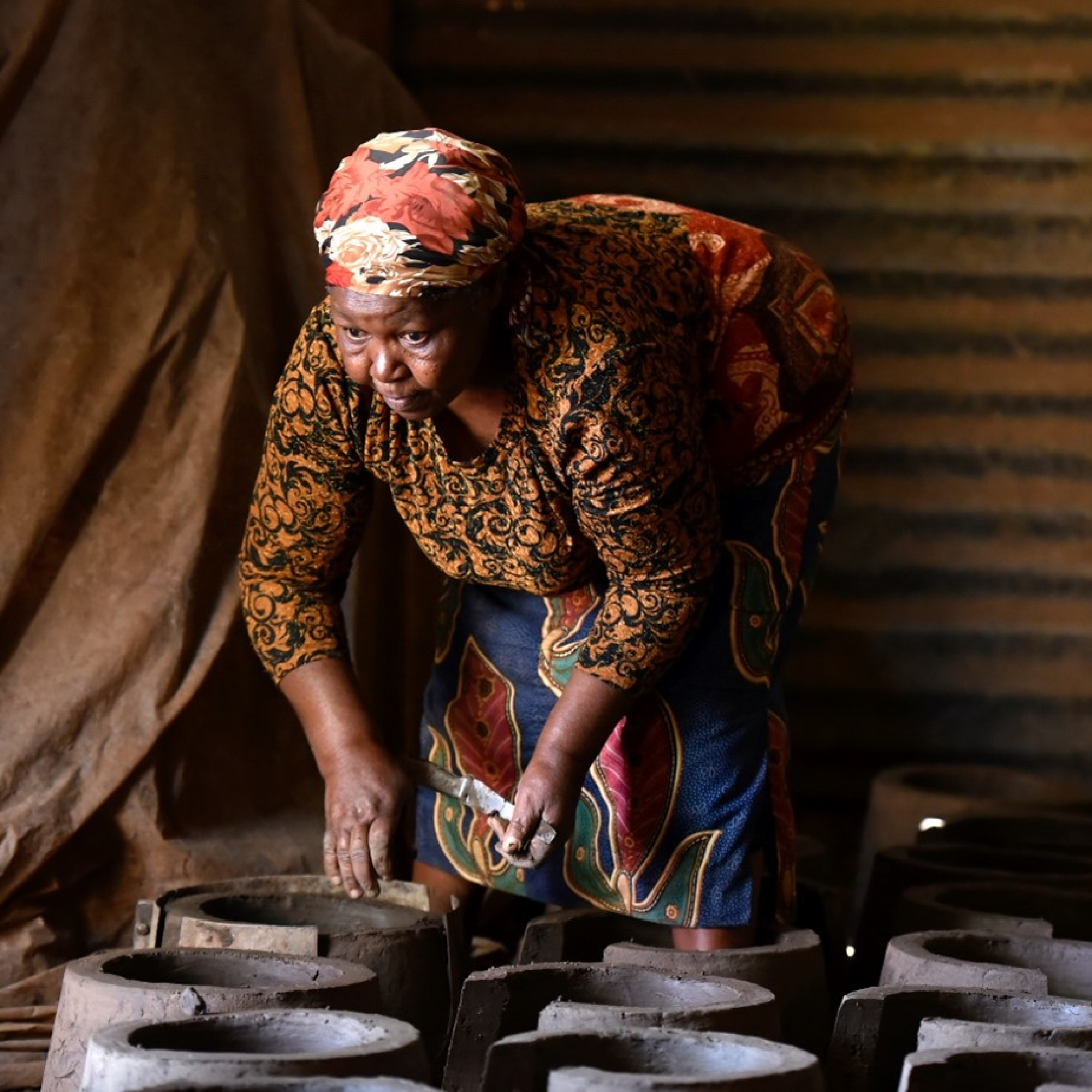 Photograph of an old black woman working with multiple cylindrical clay molds arranged on a surface inside a dimly lit space. The person wears a patterned headscarf and clothing, bending over to shape or inspect one of the molds, highlighting traditional craftsmanship or pottery-making process.