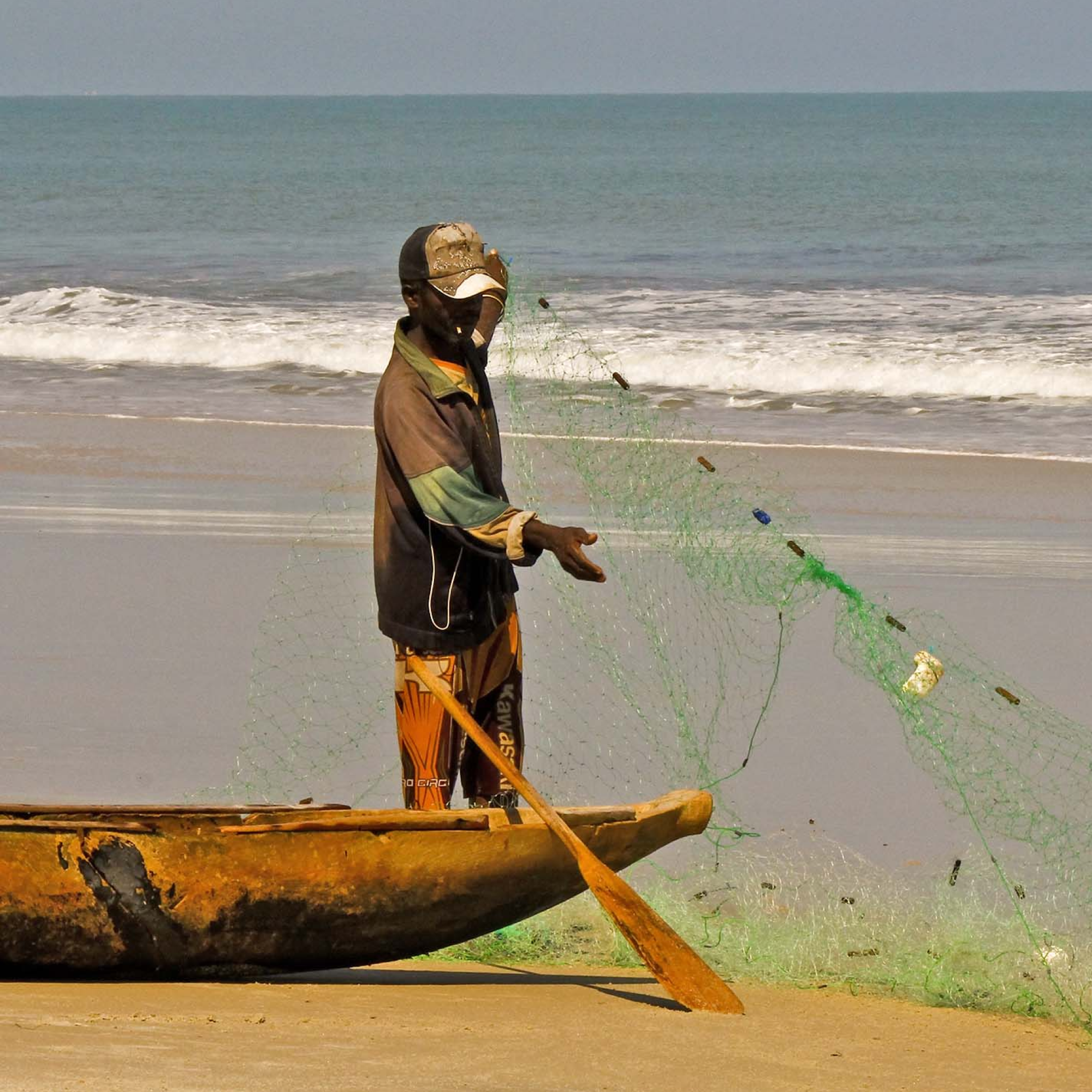 Photograph of a black fisherman standing on a sandy beach next to a wooden canoe, holding a green fishing net. Ocean waves are visible in the background, with the fisherman wearing a cap, jacket, and orange pants, preparing to cast the net.