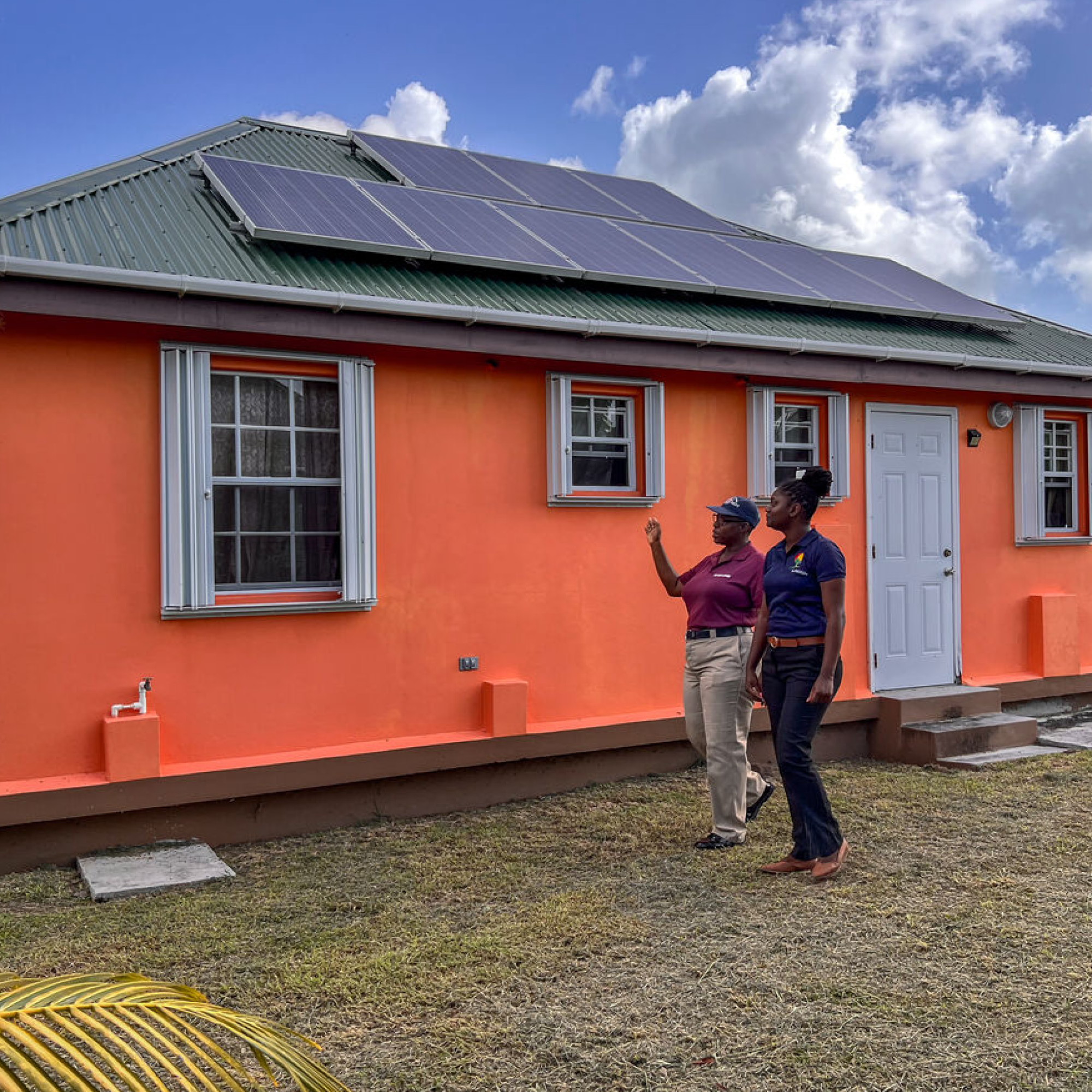Photograph showing two black women standing outside an orange house with solar panels installed on a green metal roof. The scene highlights renewable energy adoption, with the women appearing to discuss or inspect the solar panel setup under a partly cloudy sky.