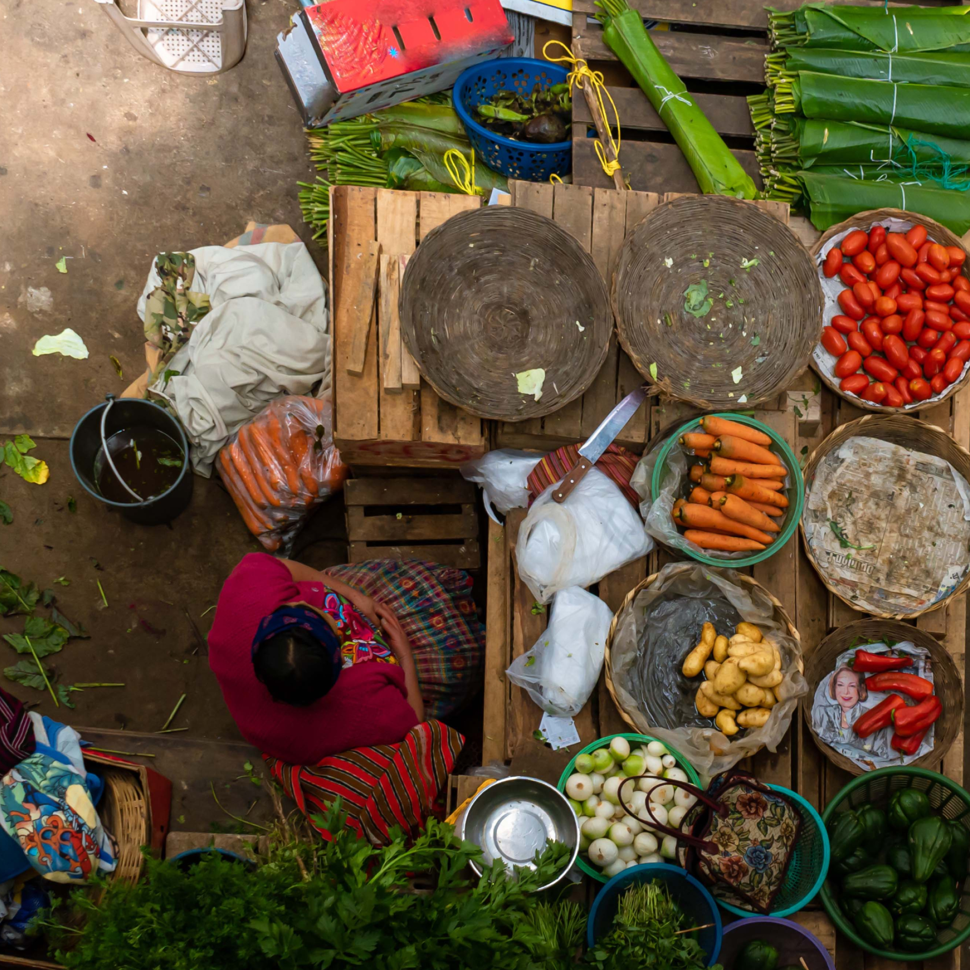 Photograph of a market stall viewed from above, displaying various fresh vegetables including carrots, tomatoes, potatoes, onions, and green peppers arranged in baskets and bowls. A vendor wearing a red top and patterned skirt is seated beside wooden crates and woven trays, with leafy greens and other produce surrounding the setup.