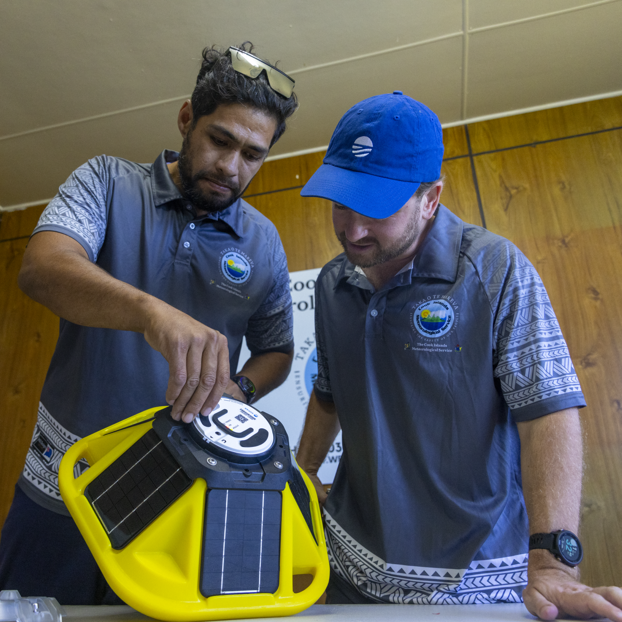 Photograph of one mixed man and one white man wearing matching gray shirts with logos, examining a yellow solar-powered device with black solar panels on a table. The setting features wood-paneled walls and a partially visible banner, suggesting a technical or environmental project demonstration.
