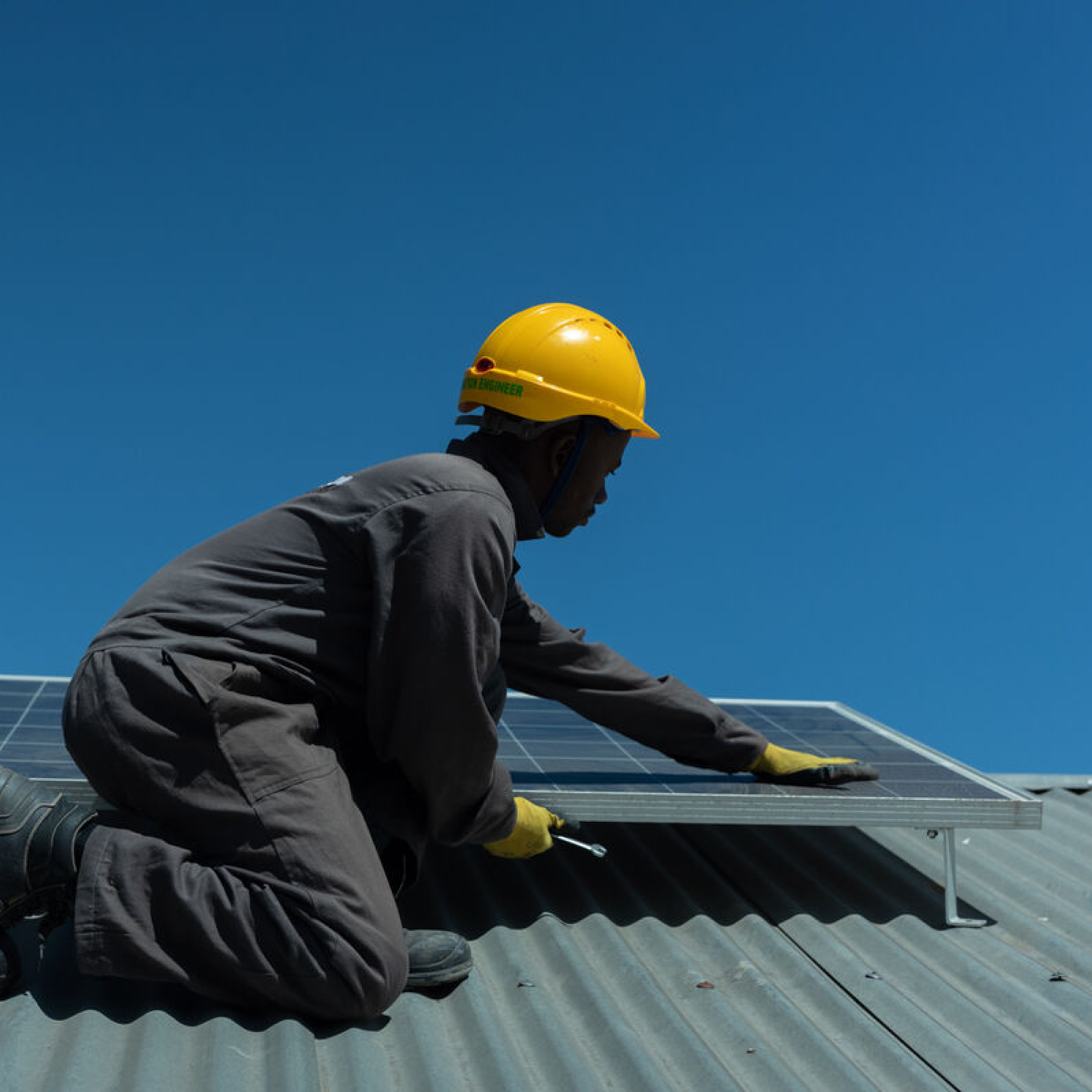 Photograph of a worker wearing a yellow hard hat and dark coveralls installing a solar panel on a corrugated metal roof under a clear blue sky. The worker is kneeling and using a tool to secure the solar panel, highlighting renewable energy installation and maintenance.
