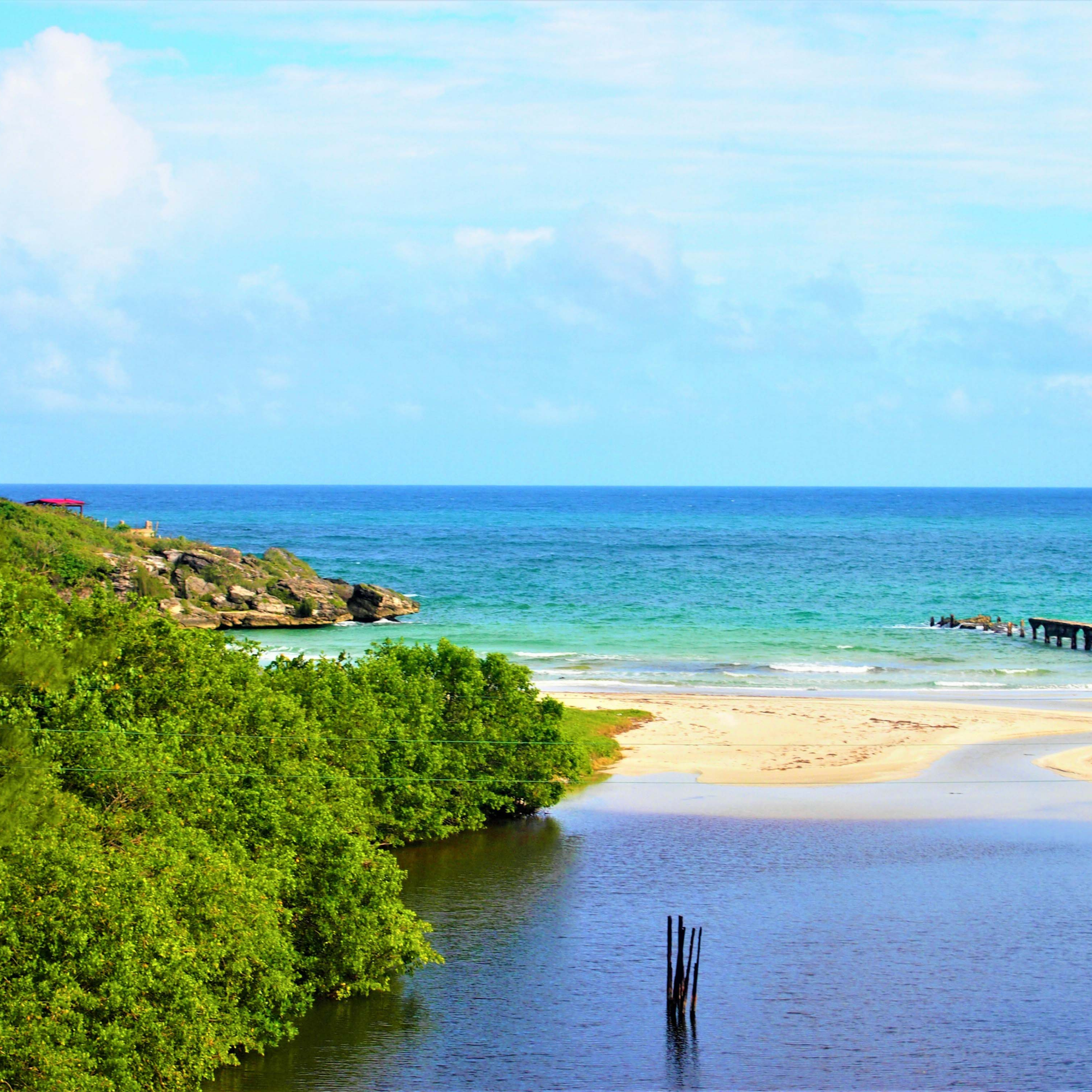 Photograph of a coastal landscape showing a sandy beach bordered by green vegetation and calm blue ocean waters under a partly cloudy sky. Key elements include a small rocky outcrop on the left, a wooden pier extending into the water on the right, and a few vertical wooden posts emerging from a shallow water area in the foreground.