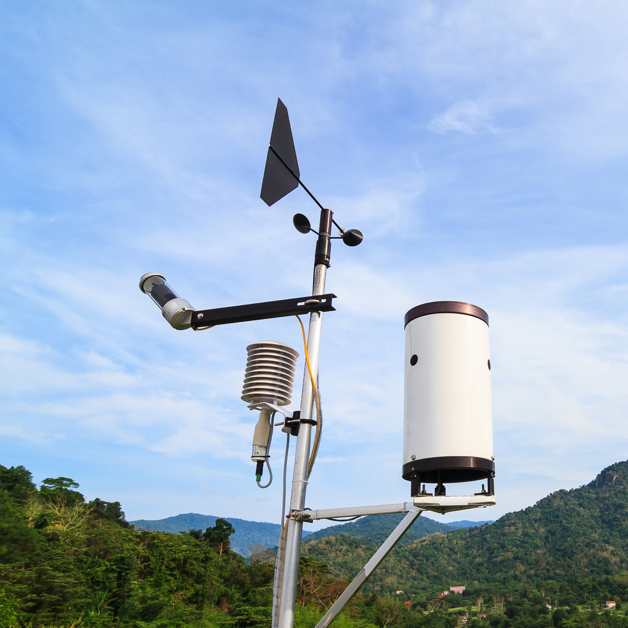 Photograph of a weather station featuring instruments including an anemometer, wind vane, rain gauge, and radiation shield mounted on a metal pole against a blue sky and forested hills background. The setup captures meteorological data such as wind speed, direction, precipitation, and temperature for environmental monitoring.