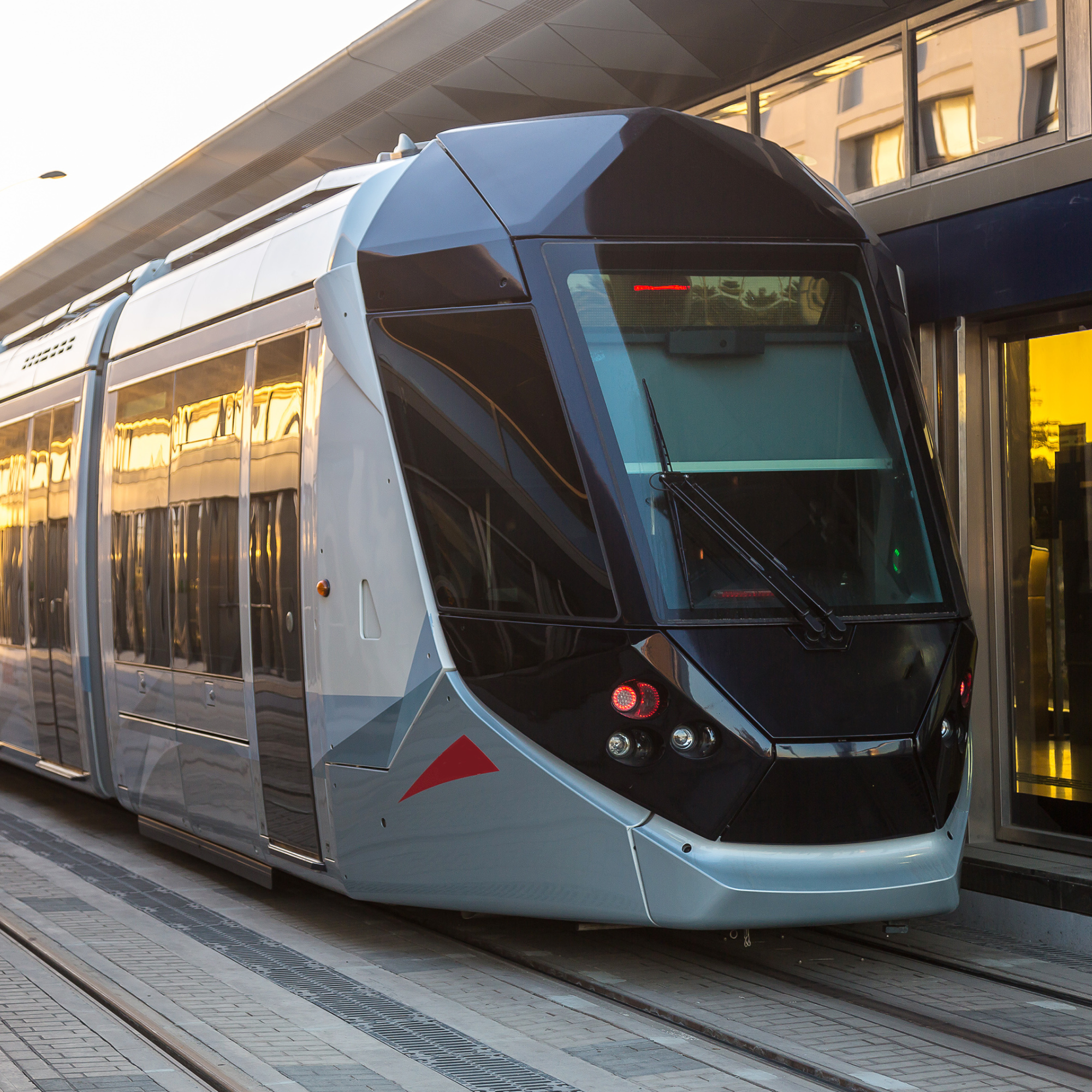 Photograph of a modern light rail train stopped at a station platform during sunset, featuring a sleek, aerodynamic design with a black front and silver body accented by a red triangular logo. Reflections on windows and station glass highlight urban surroundings, emphasizing contemporary public transit infrastructure.