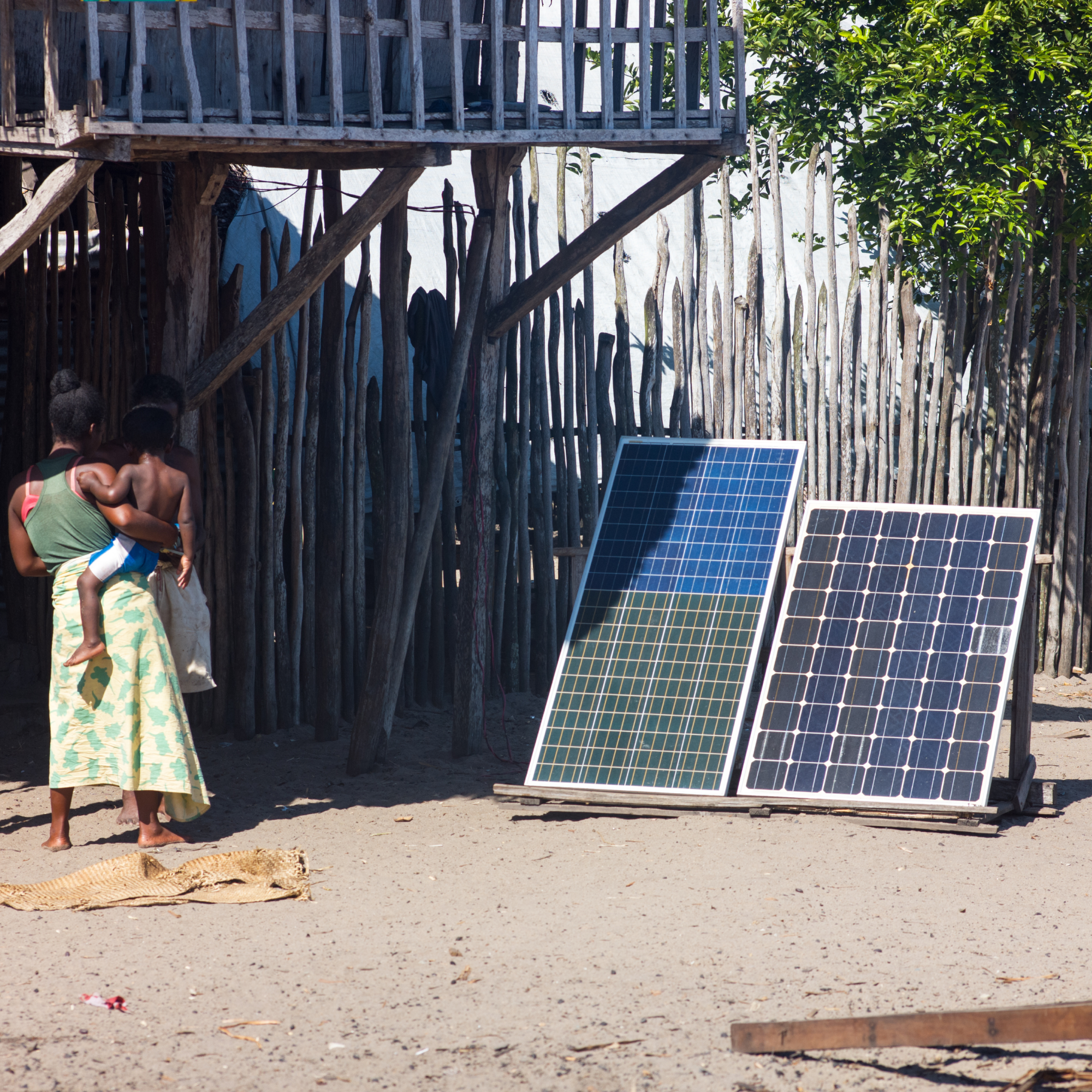 Photograph showing two solar panels mounted on a wooden frame outdoors near a rustic wooden fence and elevated structure. A black woman carrying a child and another person stand nearby, highlighting solar energy use in a rural setting.