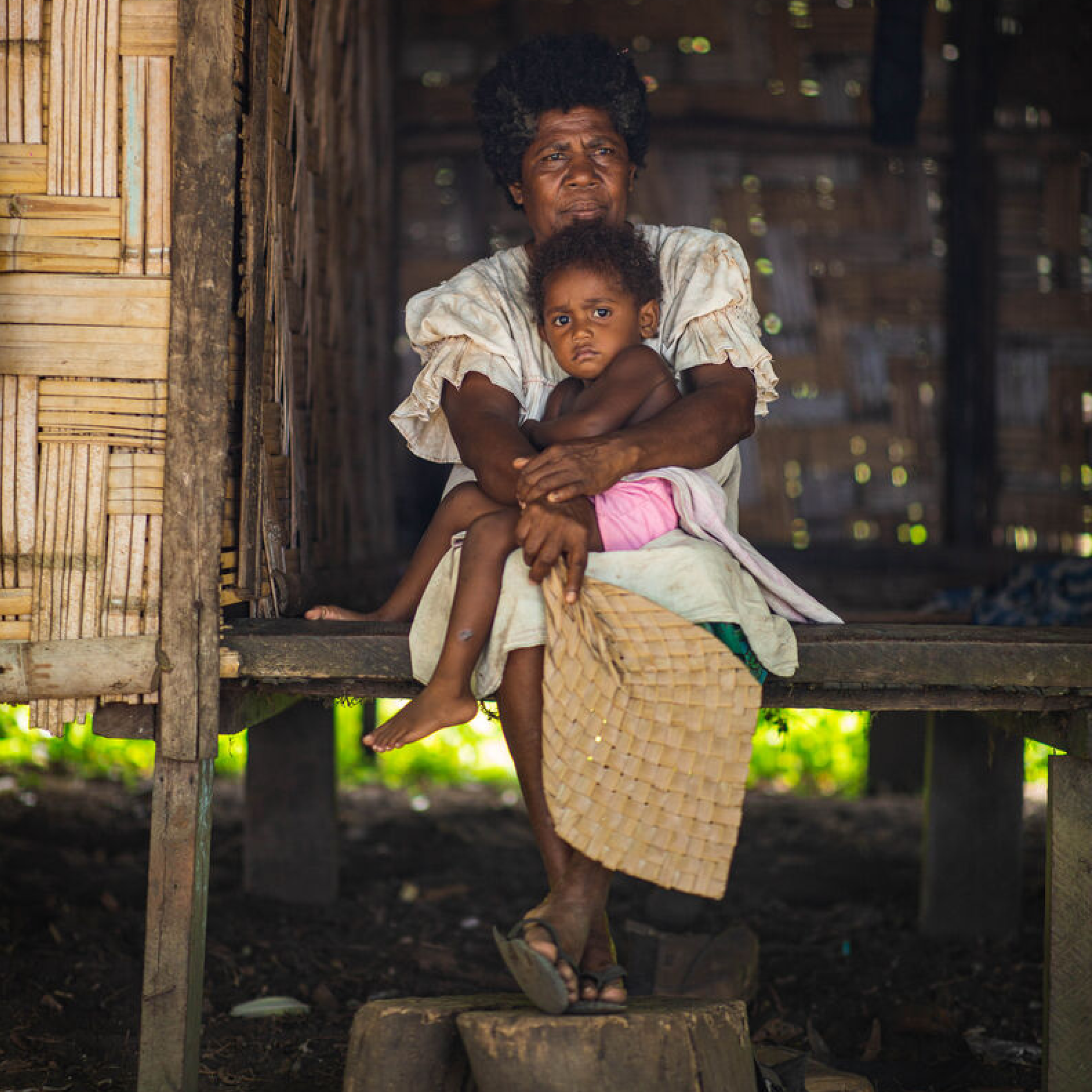 Photograph showing an old woman from Vanuatu sitting on a wooden platform inside a rustic structure, holding a child on their lap. The setting features natural light filtering through gaps in woven walls, highlighting traditional clothing and a woven mat held by the person.