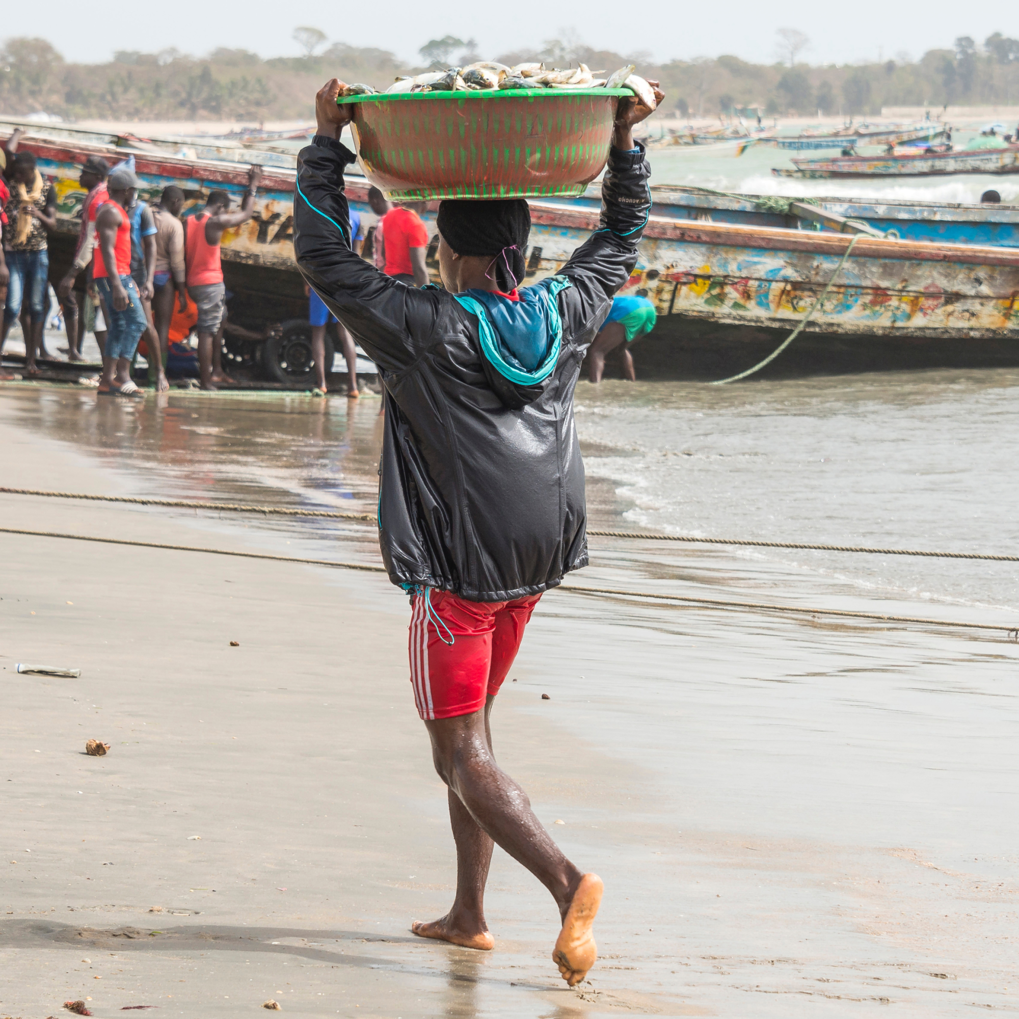 Photograph of a black young man walking barefoot on a sandy shore carrying a large basket of fish on their head, with several boats and people in the background engaged in fishing activities. The scene captures a coastal fishing community, highlighting daily labor and traditional fish harvesting methods.