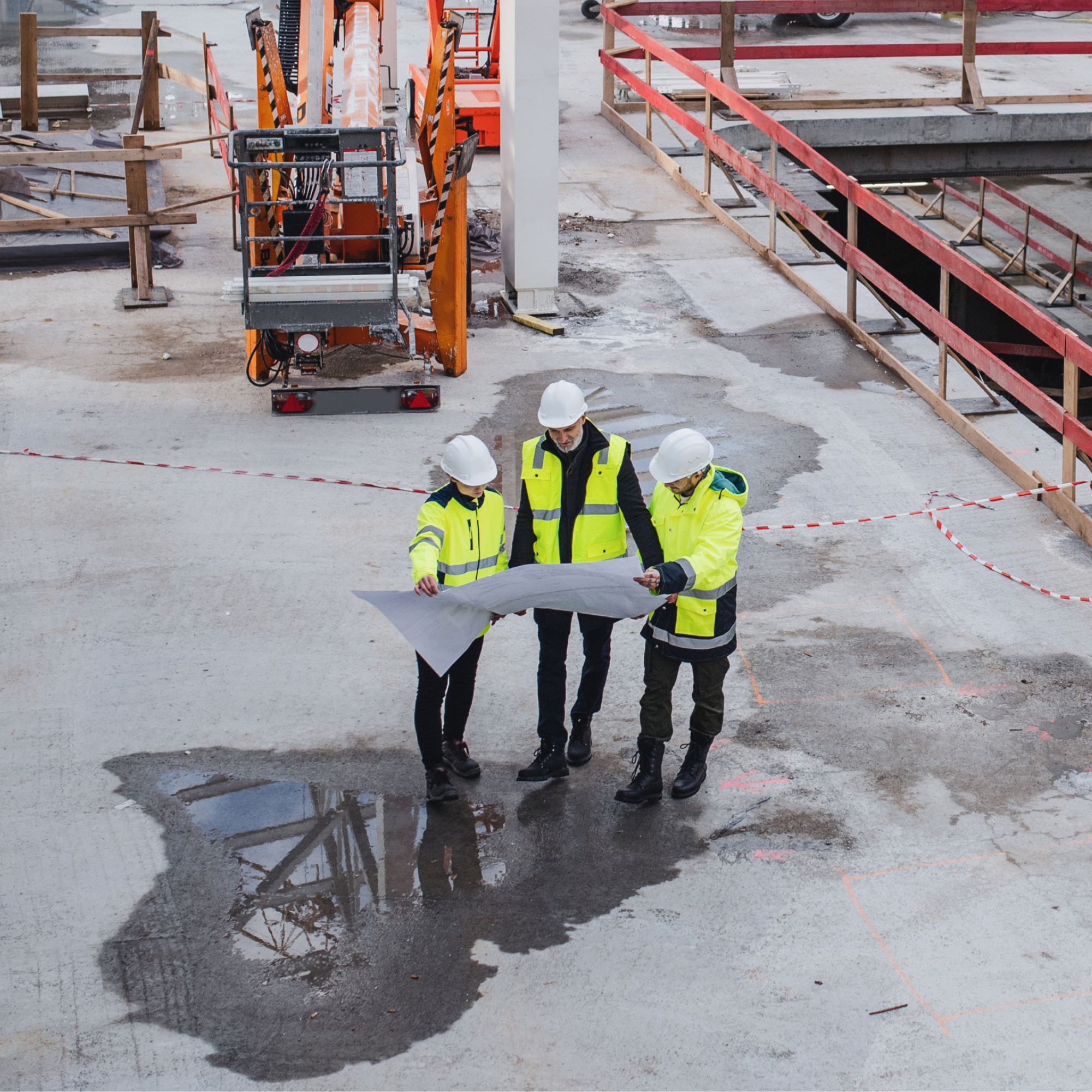Photograph showing three construction workers wearing high-visibility jackets and white helmets reviewing blueprints on a concrete site with safety barriers and construction equipment in the background. The scene highlights collaboration and planning during an active building project with visible water puddles and marked areas on the ground.