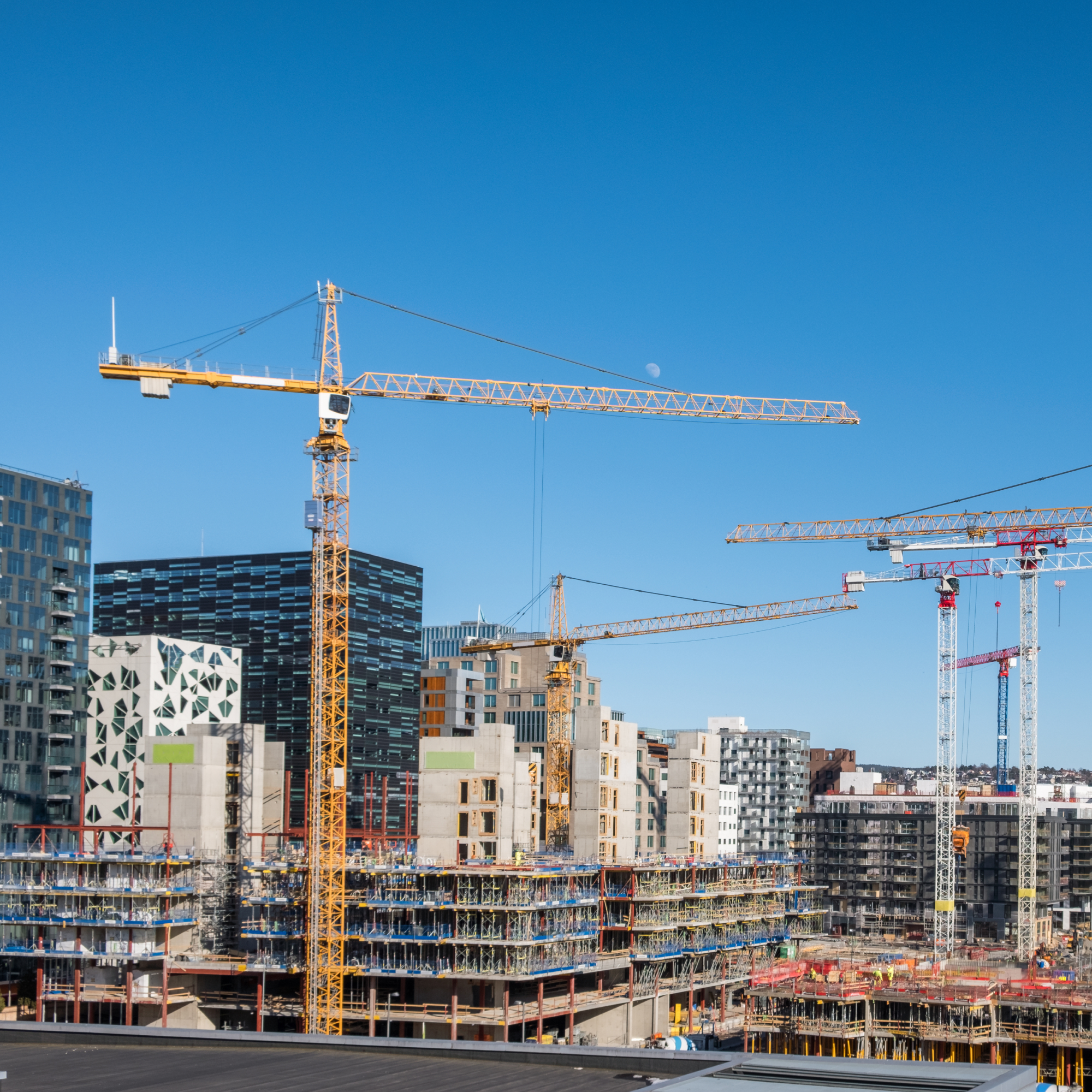 Photograph of an urban construction site featuring multiple yellow and white tower cranes actively working on several multi-story buildings in various stages of development. Modern high-rise buildings with distinct geometric patterns and glass facades form the backdrop under a clear blue sky, highlighting ongoing city expansion and infrastructure growth.