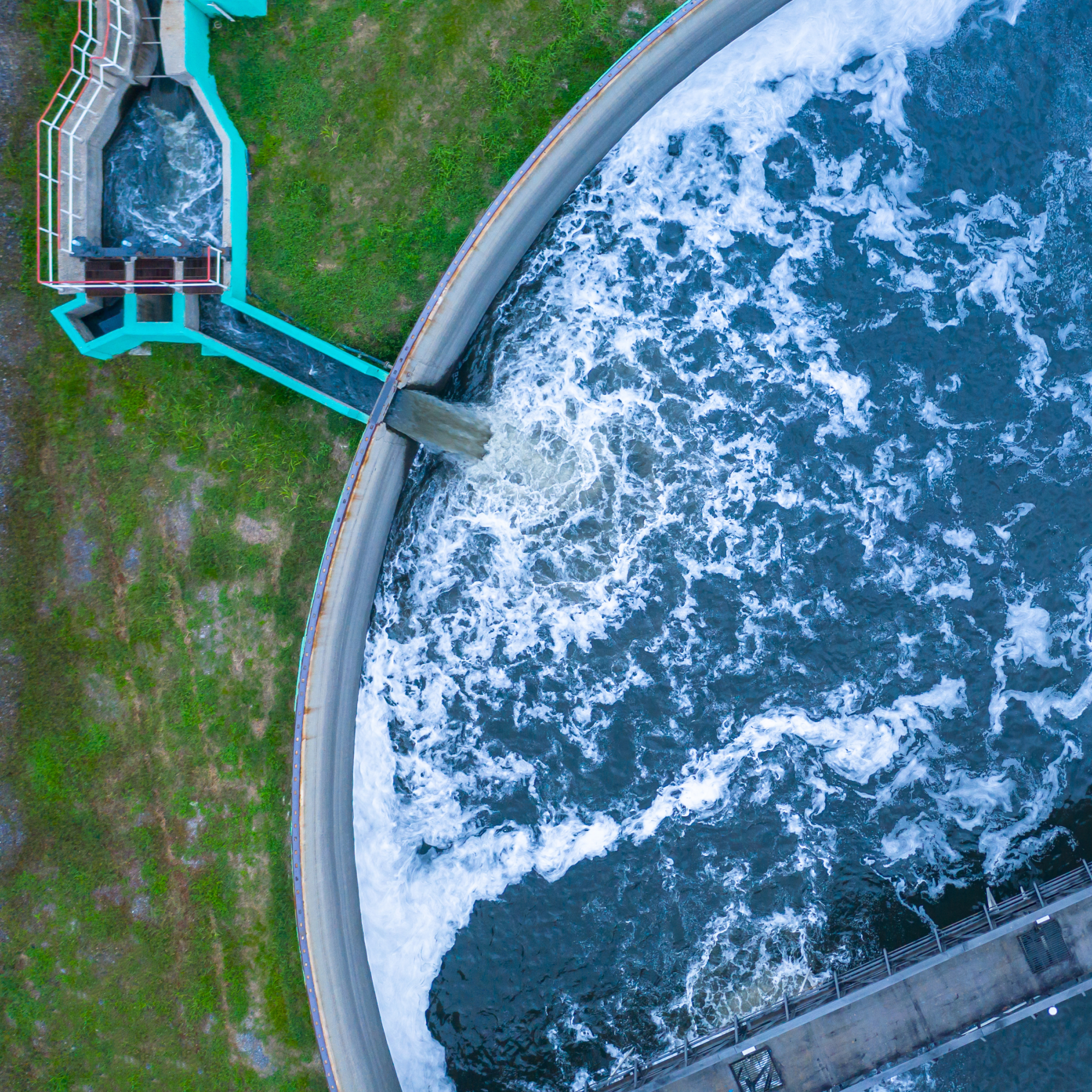Overhead view of a circular aeration basin at a water treatment plant. Wastewater flows from a teal concrete channel into the large tank, creating white foam and turbulent waves as it undergoes purification. A metal access bridge is visible in the lower right.
