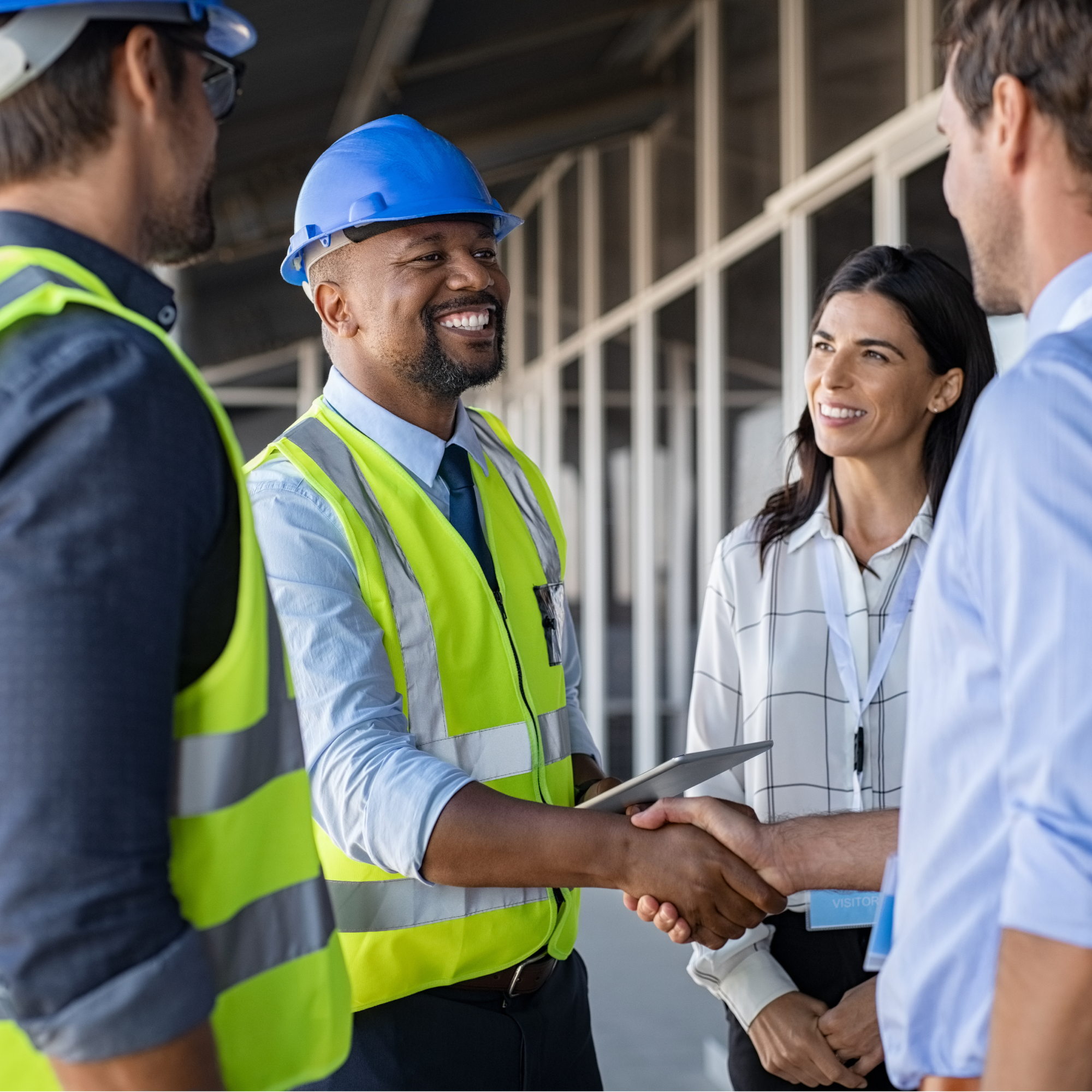 Photograph showing four professionals in a construction or industrial setting, with two men shaking hands in the foreground. Two individuals wear safety vests and helmets, while the other two wear business attire, indicating a meeting or agreement between construction and office teams.