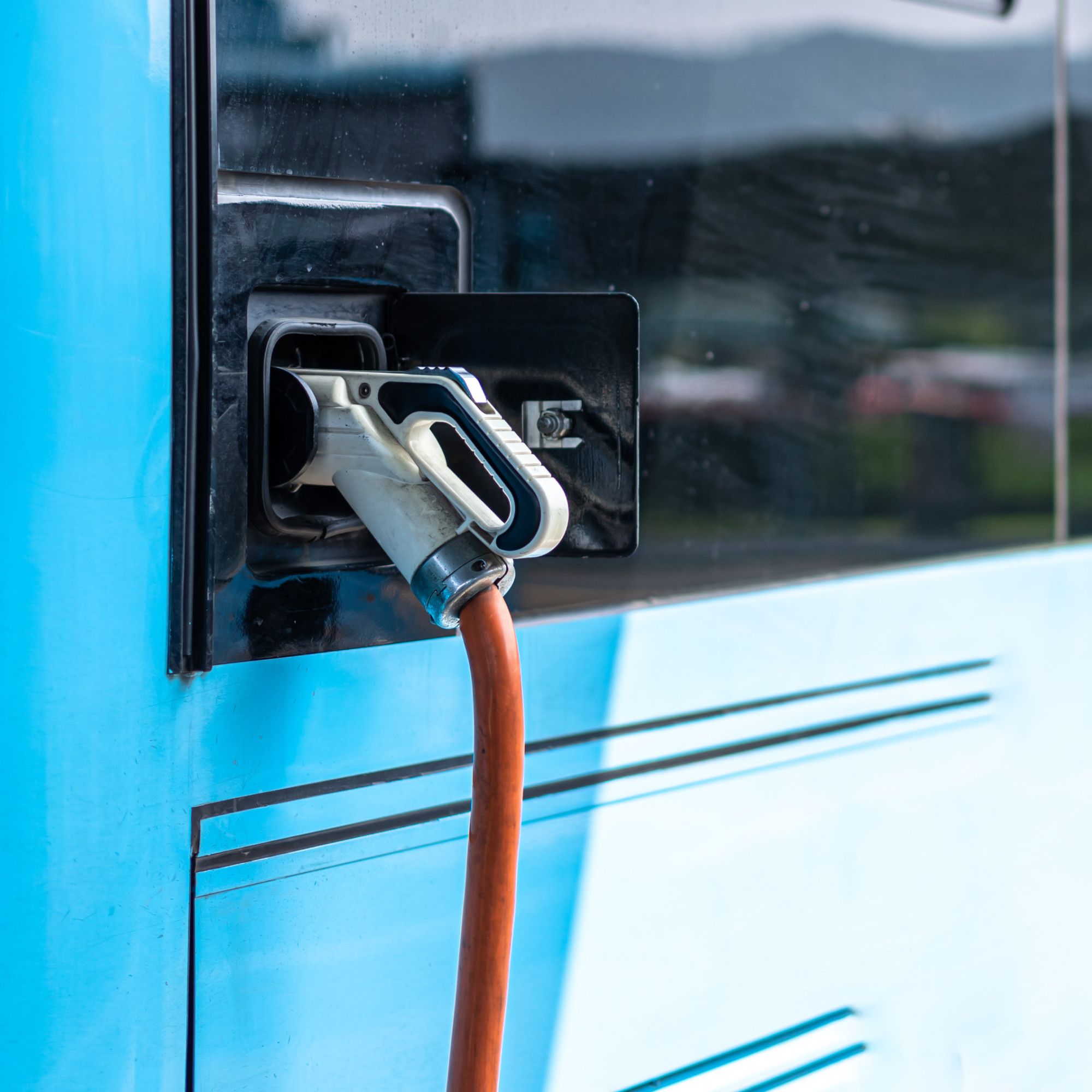 Photograph showing a close-up of an electric vehicle charging port with a white and black charging connector plugged in, attached to an orange cable. The vehicle is light blue with black accents, highlighting the charging process and promoting electric mobility.