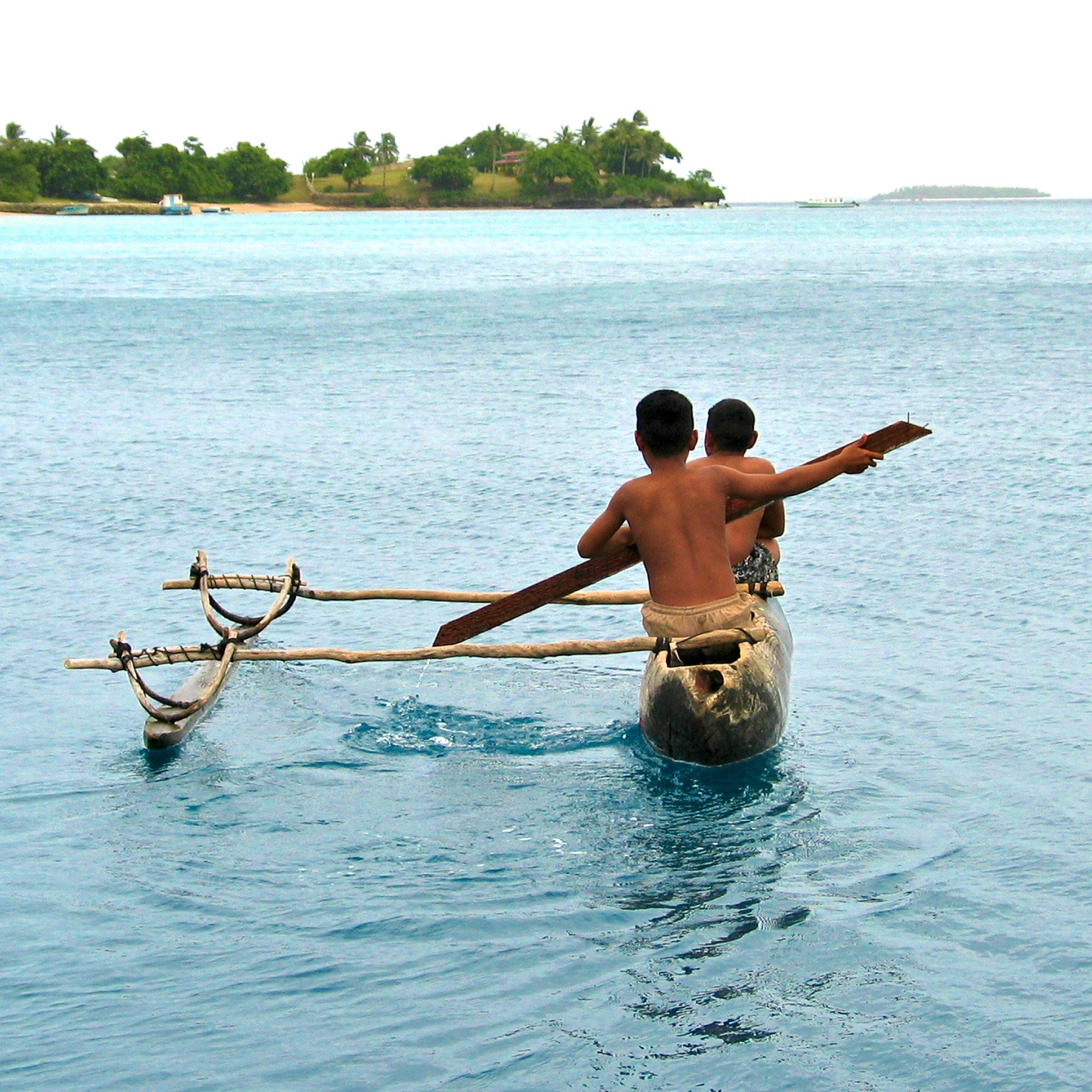 Photograph of two boys paddling a traditional outrigger canoe on calm blue water near a small island with palm trees. The canoe features wooden outriggers on both sides for stability, and the boys are shirtless, focusing on navigating toward the island in the distance.