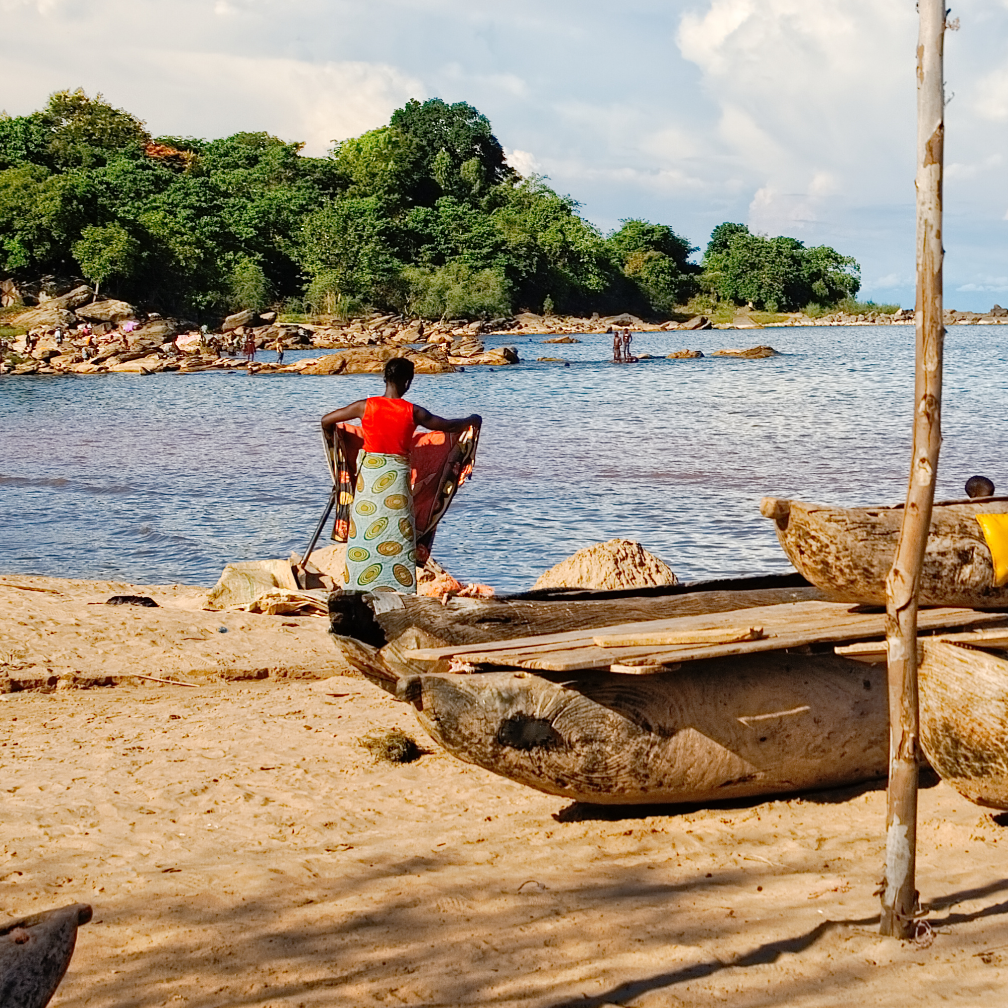 Photograph of a lakeside scene showing a woman in a red top and patterned skirt standing on sandy shore near wooden boats, with lush green trees and rocky shoreline in background. The setting highlights daily life by water, featuring natural elements like water, rocks, and vegetation under a partly cloudy sky.