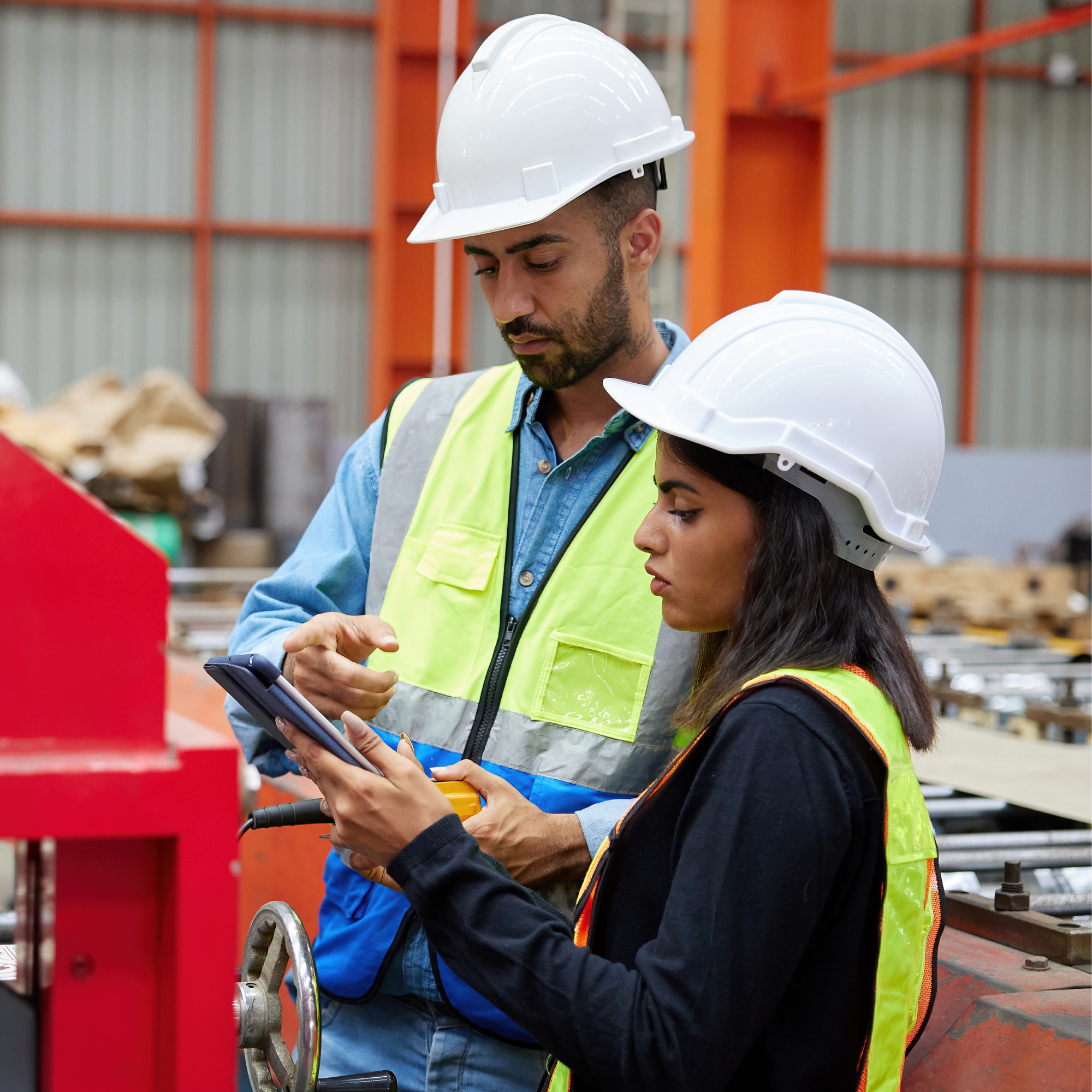 Photograph of two factory workers wearing white hard hats and safety vests, discussing information on a tablet inside an industrial facility. Background includes machinery and metal structures, highlighting a collaborative work environment focused on manufacturing or production processes.