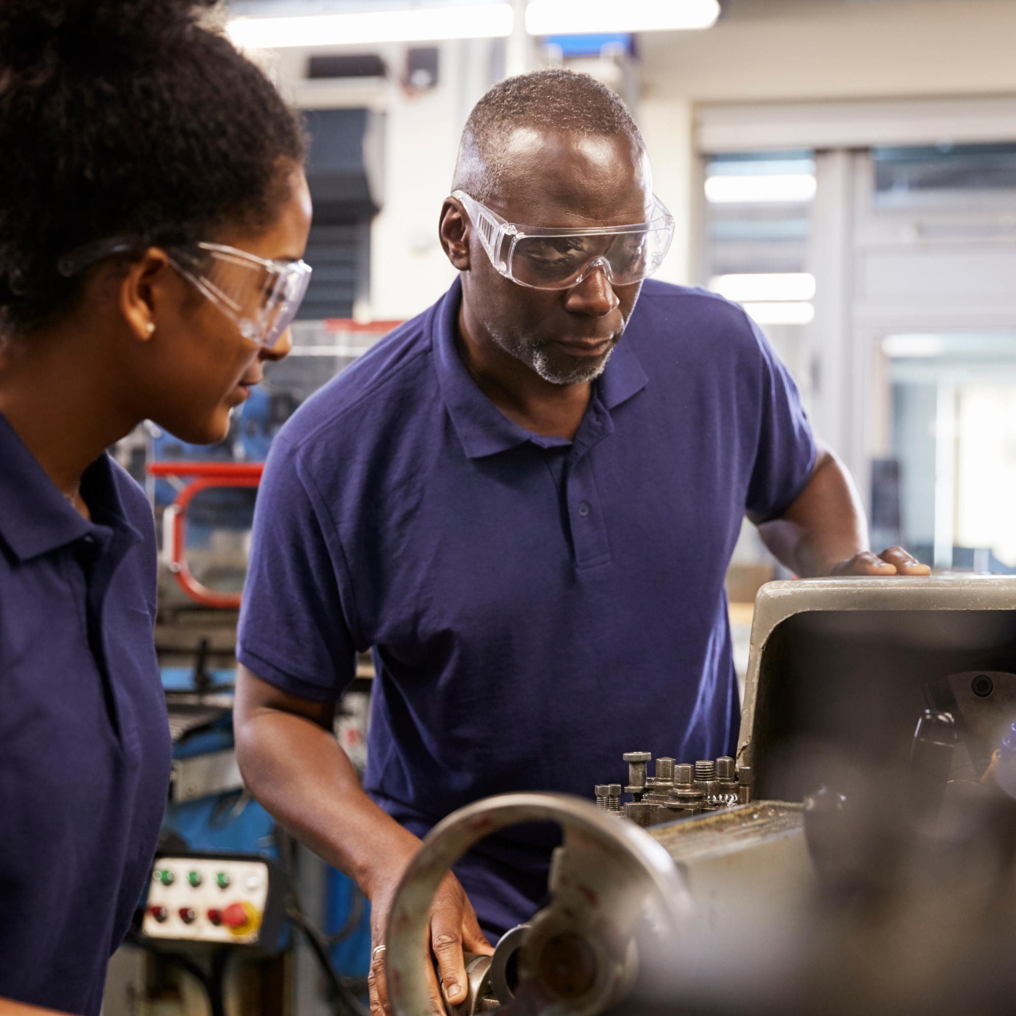 Photograph of a black woman and a black man wearing blue purple polo shirts working with industrial machinery in a workshop setting. One person is closely inspecting or adjusting a metal component on the machine while the other observes, with various tools and equipment visible in the background.