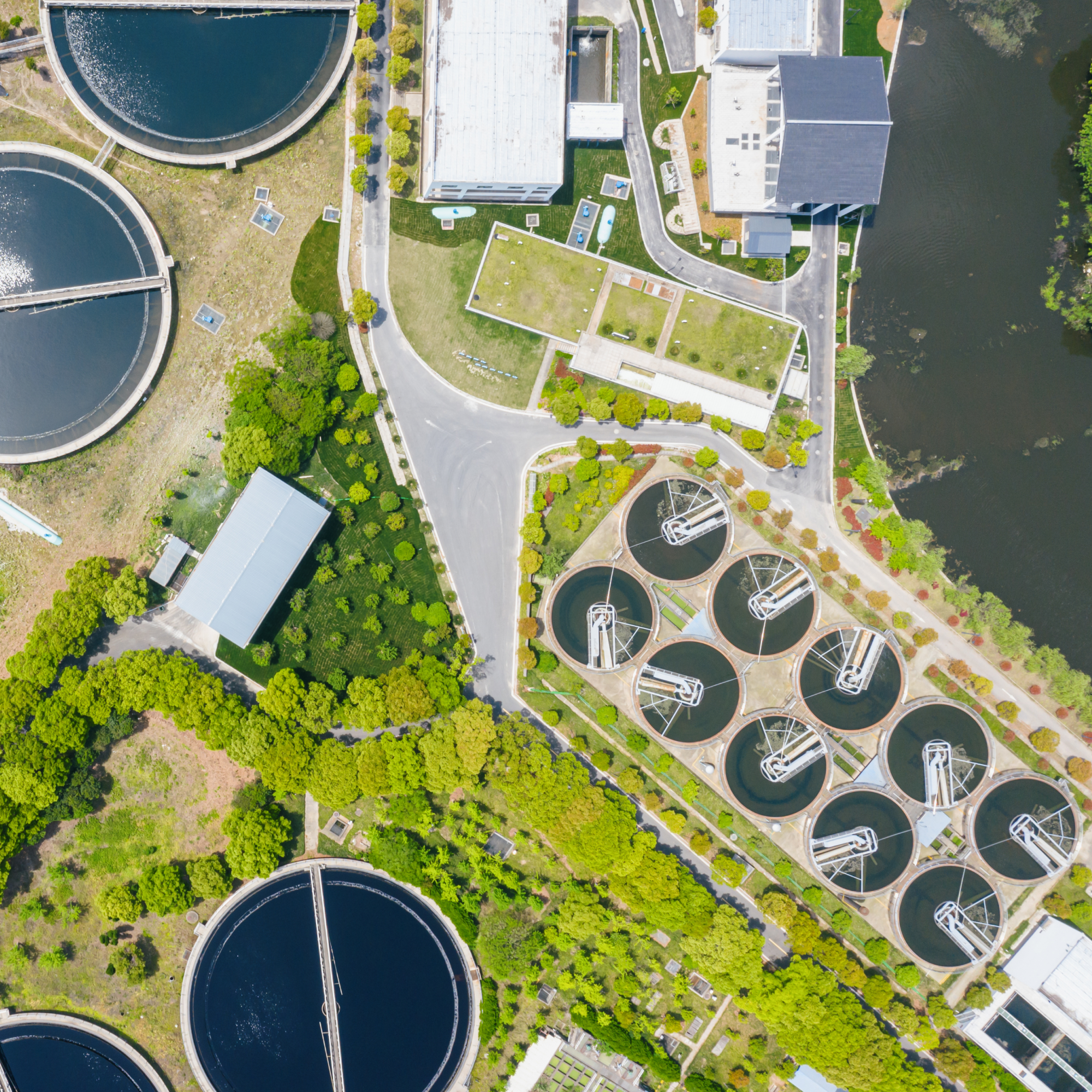 Aerial photograph of a wastewater treatment plant showing multiple circular clarifier tanks and rectangular buildings surrounded by greenery and roads. The layout highlights key structural components such as aeration basins, sedimentation tanks, and administrative buildings, illustrating the facility's water purification process.