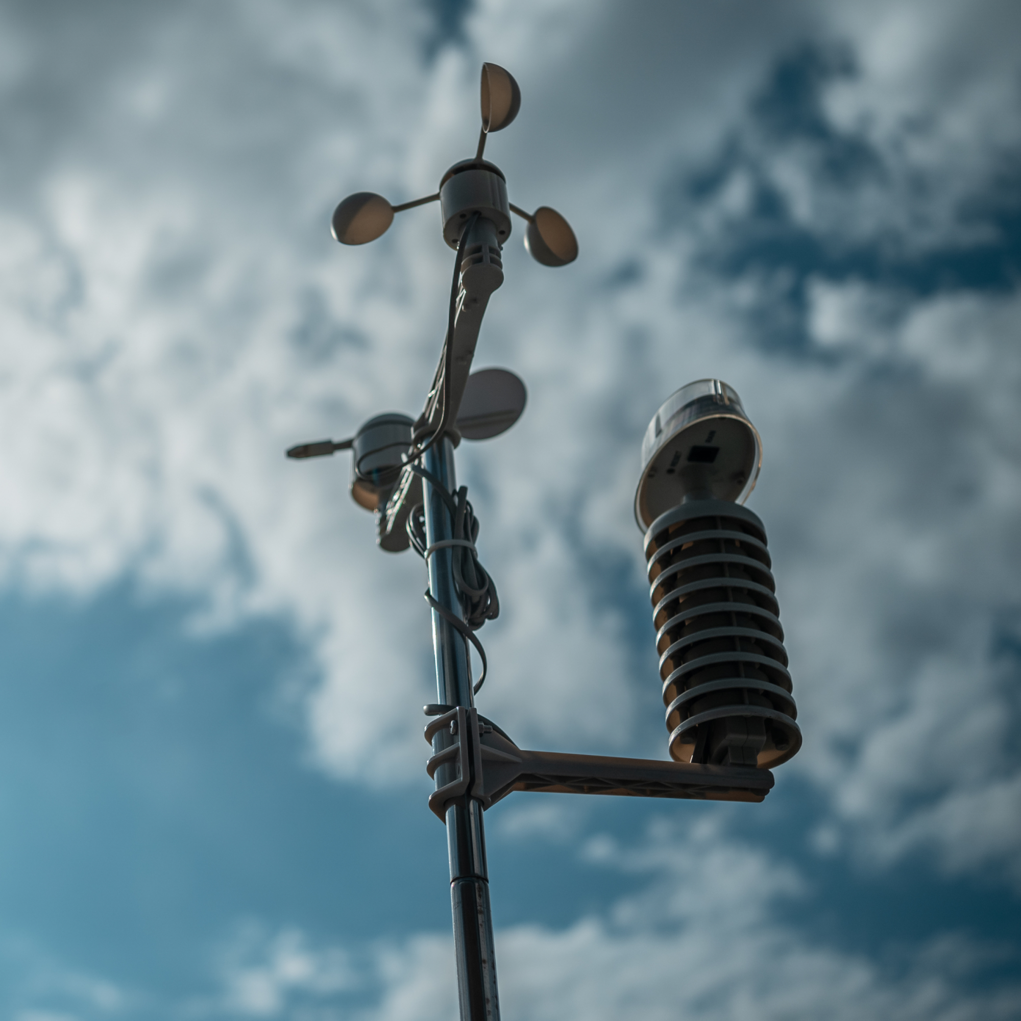 Photograph of a weather station mounted on a pole against a cloudy blue sky, featuring an anemometer with three cups for measuring wind speed and a cylindrical radiation shield housing temperature and humidity sensors. The setup includes visible wiring and a bracket supporting the radiation shield, highlighting components used for meteorological data collection.