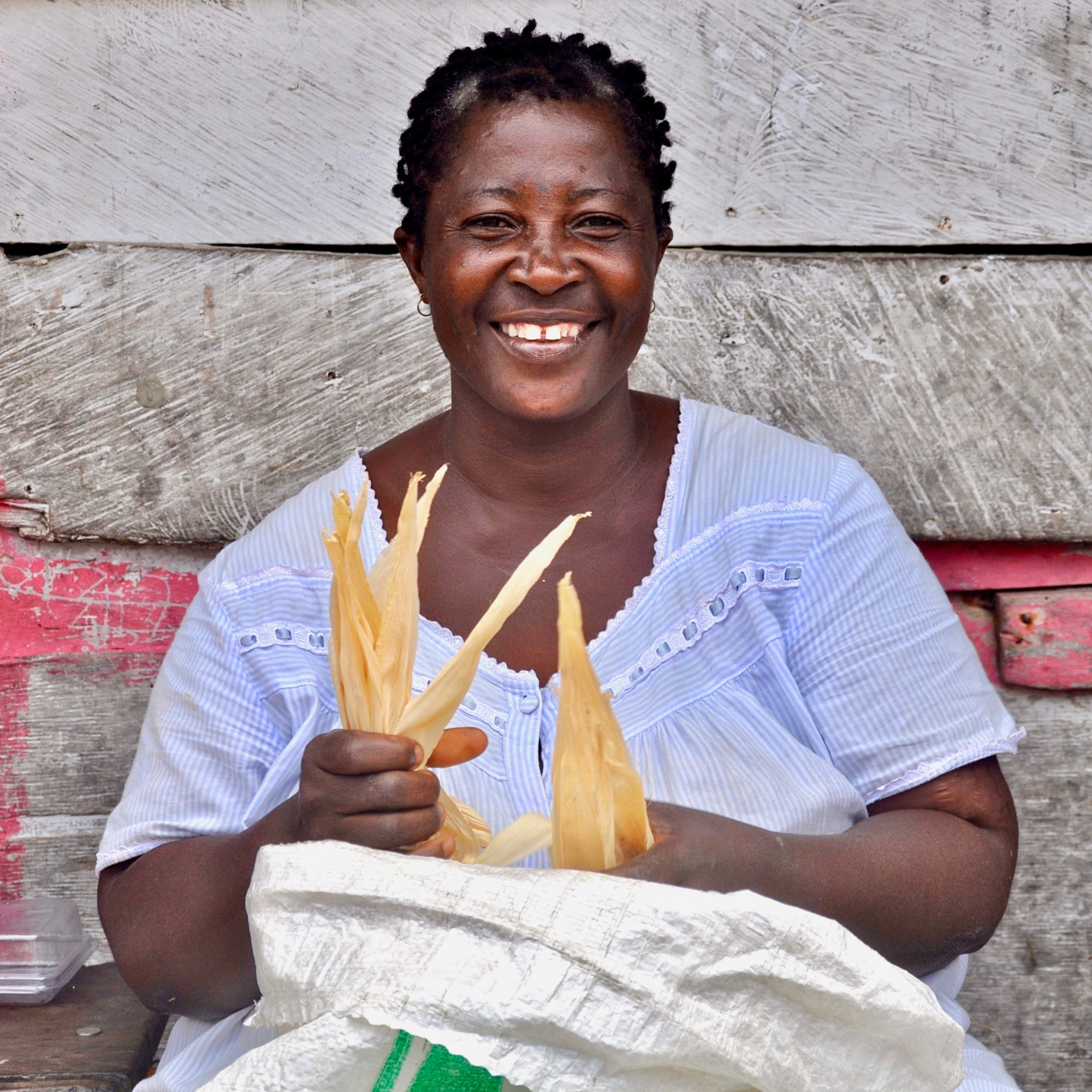 Photograph of a person wearing a white blouse sitting against a wooden wall while holding peeled corn husks. The scene highlights traditional food preparation with rustic background elements including weathered wood and a white sack.