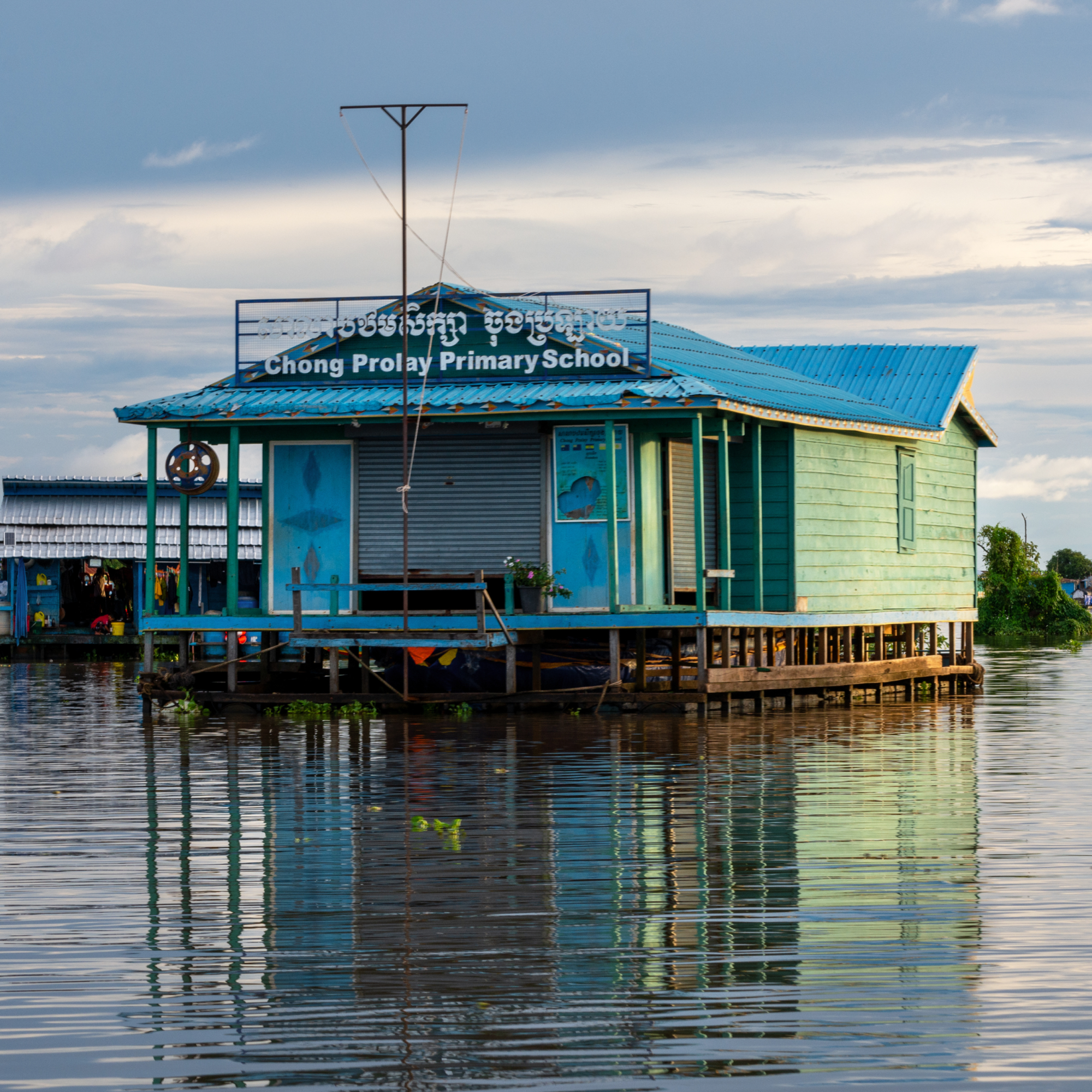 Photograph of a floating primary school building named Chong Prolay Primary School, situated on stilts above water with a blue roof and green wooden walls. The structure reflects in calm water under a partly cloudy sky, highlighting adaptation to a flood-prone or aquatic environment.