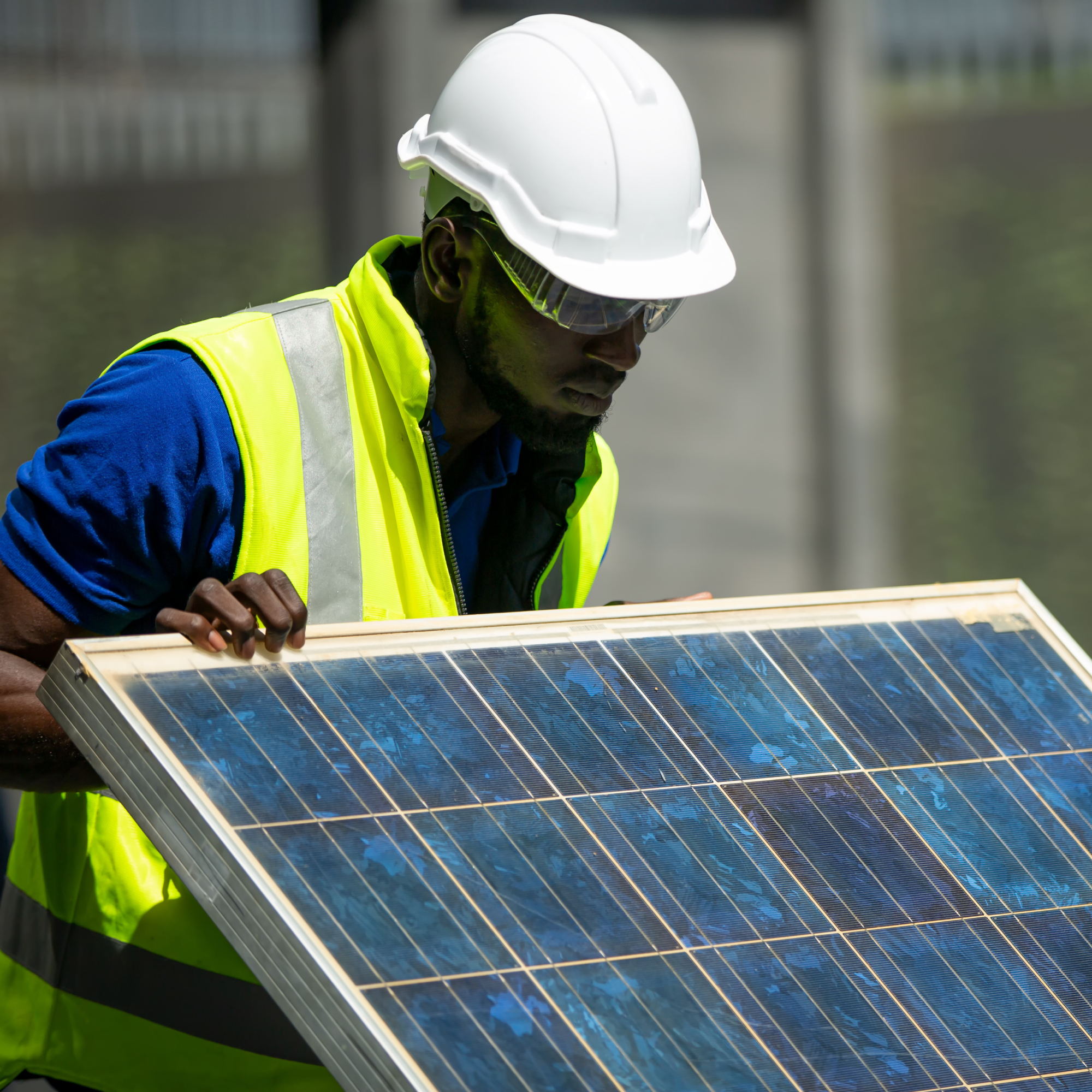 Photograph of a construction worker wearing a white hard hat and yellow safety vest holding a blue solar panel. The scene highlights renewable energy installation with focus on worker's careful handling of solar technology.