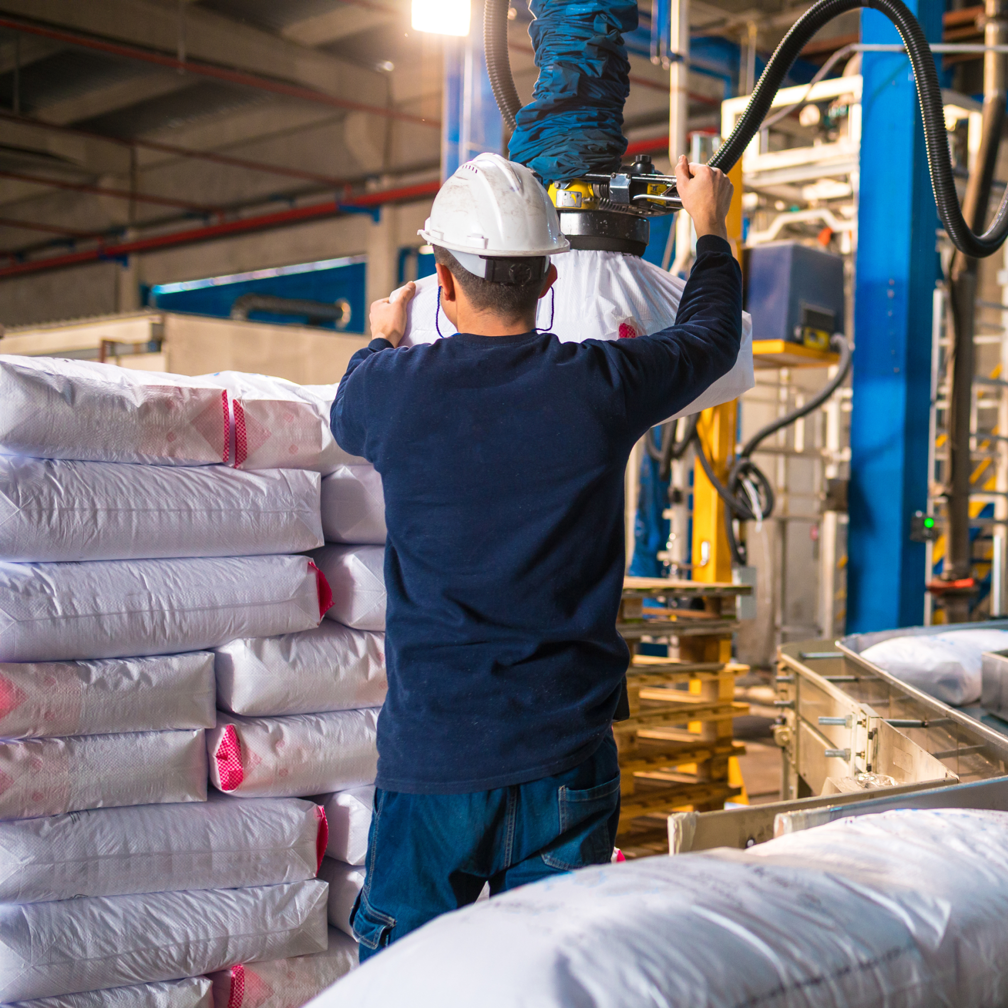 Photograph of a factory worker wearing a white hard hat and dark clothing operating machinery to lift and move large white bags stacked on pallets. The industrial setting includes metal structures, conveyor belts, and equipment, highlighting a packaging or material handling process.