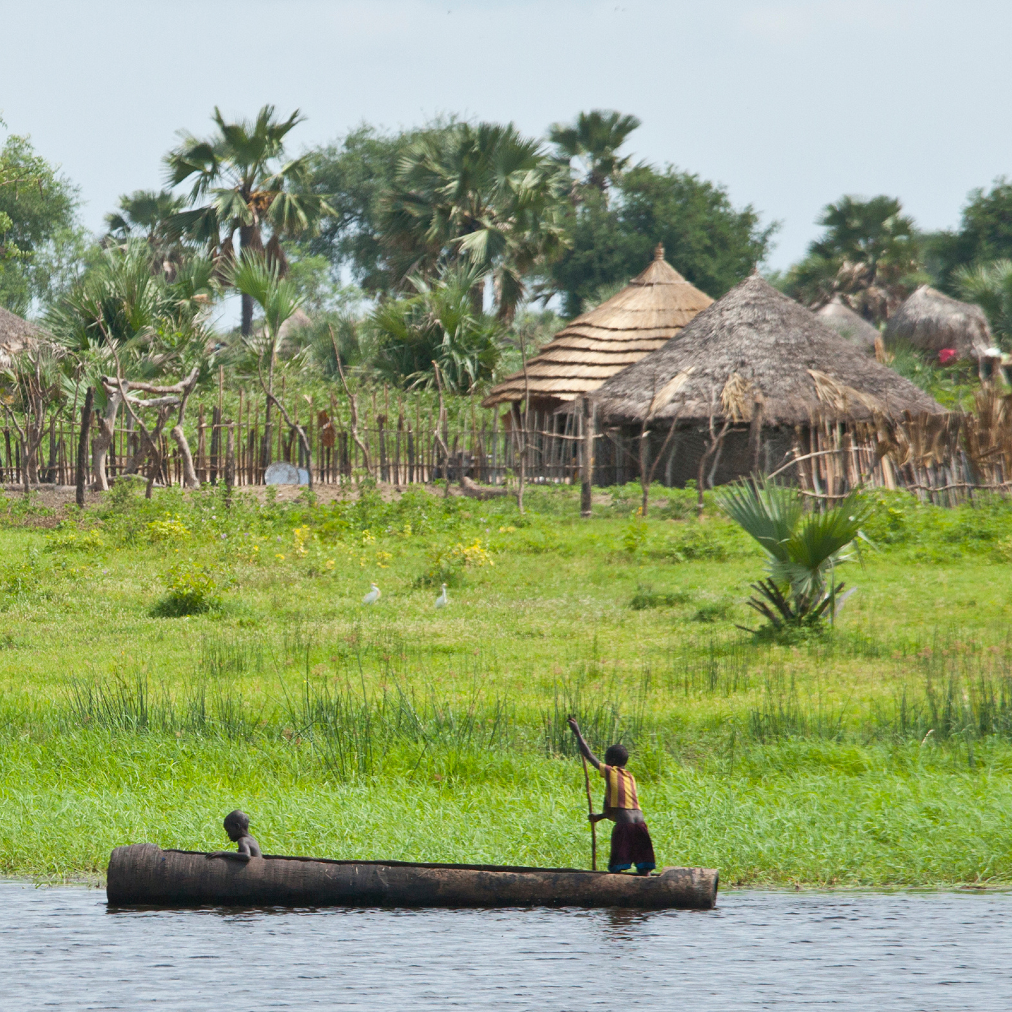 Photograph showing two people in a wooden canoe on a body of water with a village featuring traditional thatched huts and palm trees in the background. The scene highlights rural life and local transportation methods in a lush, green environment.