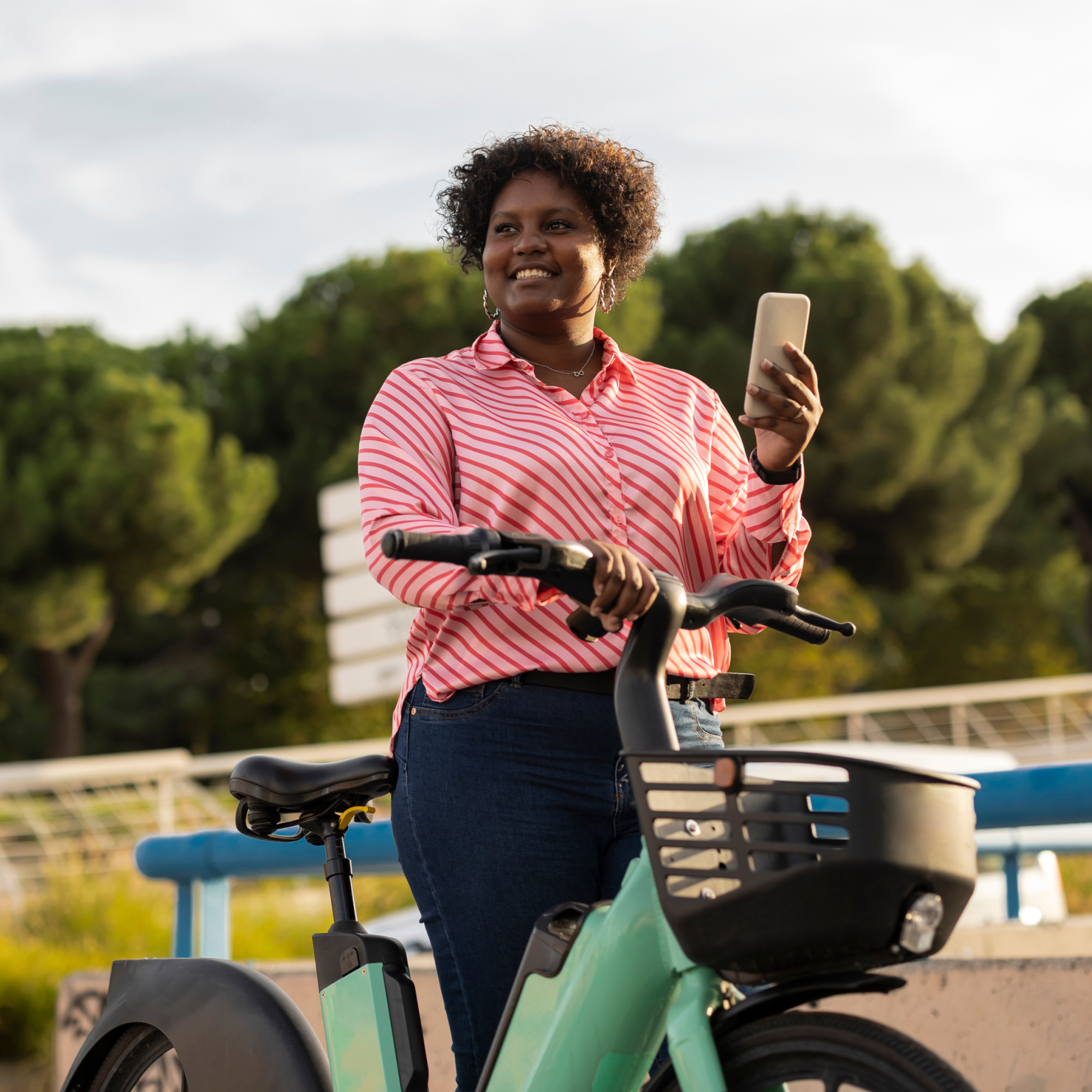 Photograph of a person standing outdoors next to a teal electric bike, holding a smartphone in one hand and gripping the bike's handlebar with the other. The person wears a pink and white striped shirt and blue jeans, with green trees and a blue railing visible in the background.